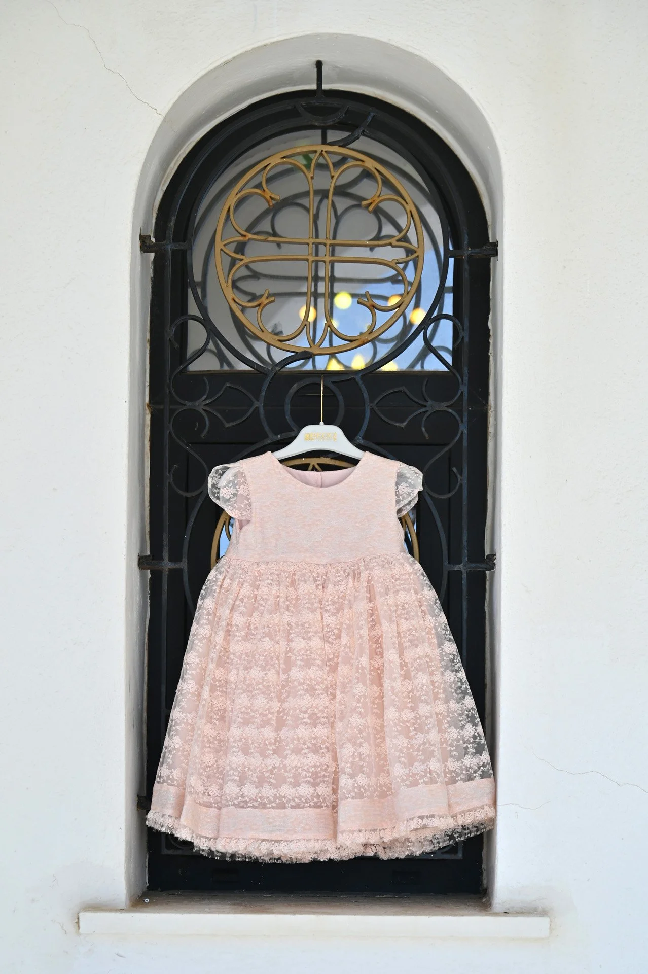 A pink, lace dress hanging on a white hanger inside a black wrought-iron and glass decorative window.