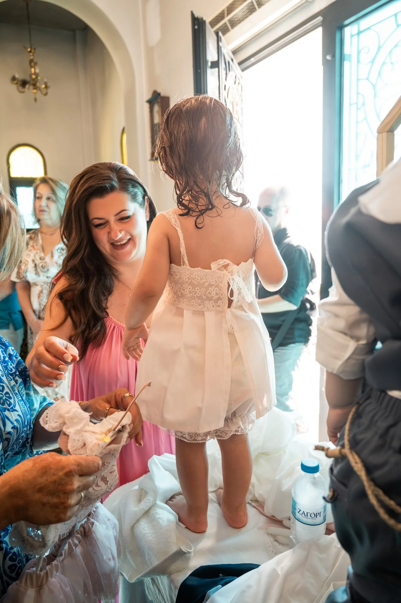 A group of women and a young girl dressed in white lingerie at a celebration, possibly a wedding, with sunlight shining through an open door in the background.
