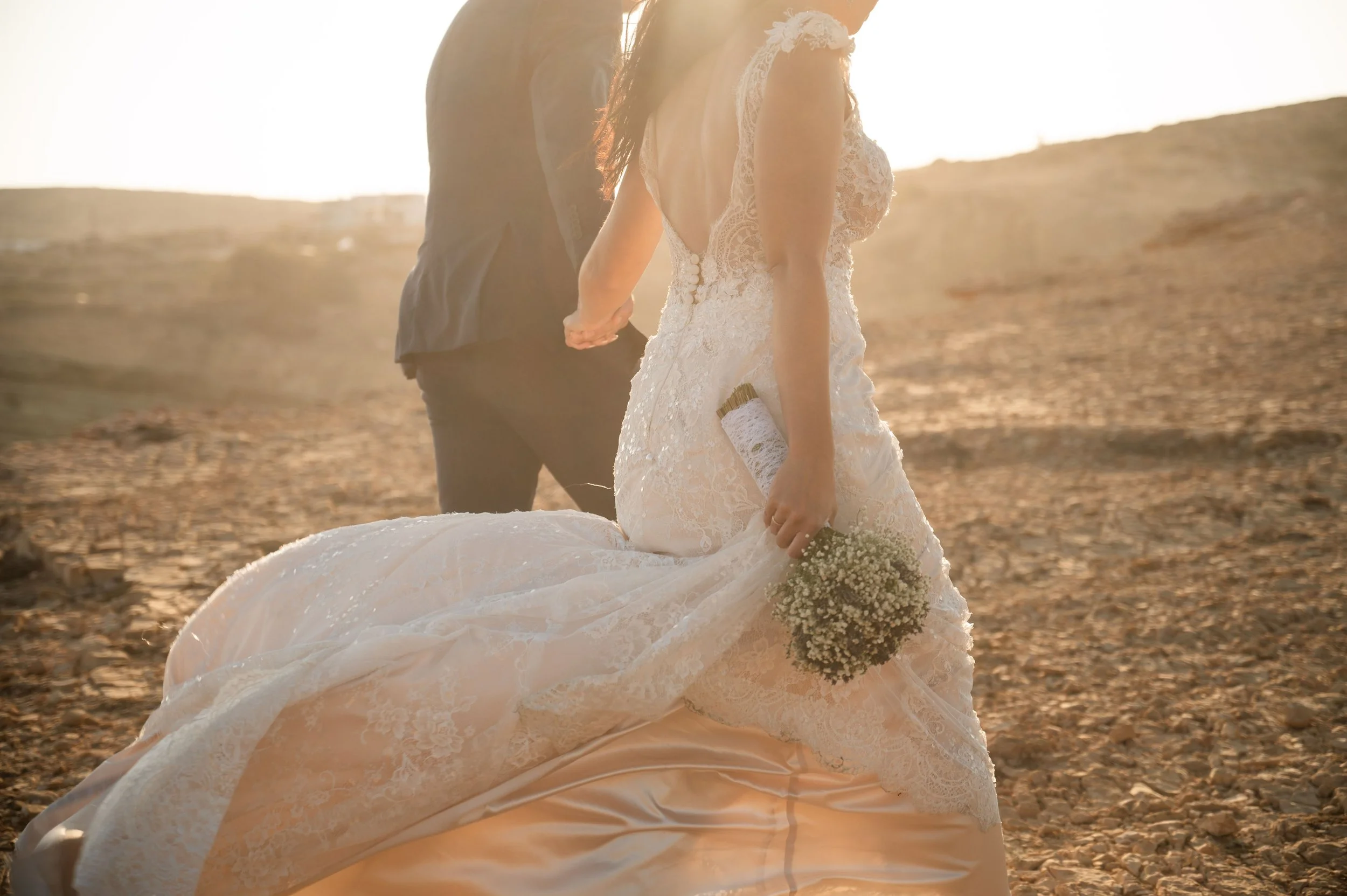 Bride in a lace wedding dress holding a bouquet of baby's breath flowers, standing in a barren outdoor landscape during sunset with groom behind her.