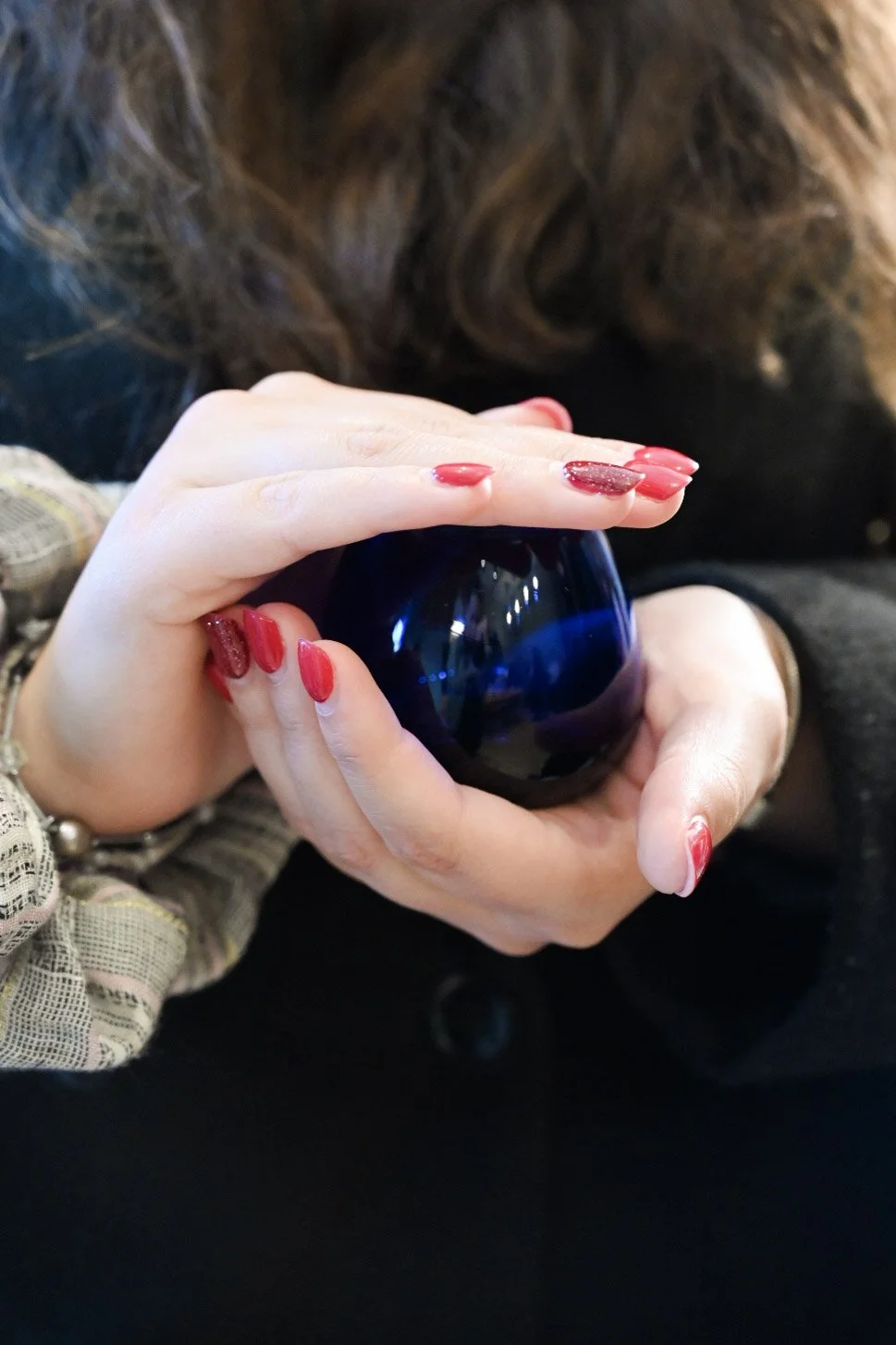 A person holding a dark blue glass candle holder with both hands, with long red painted nails, and a person with brown hair in the background.