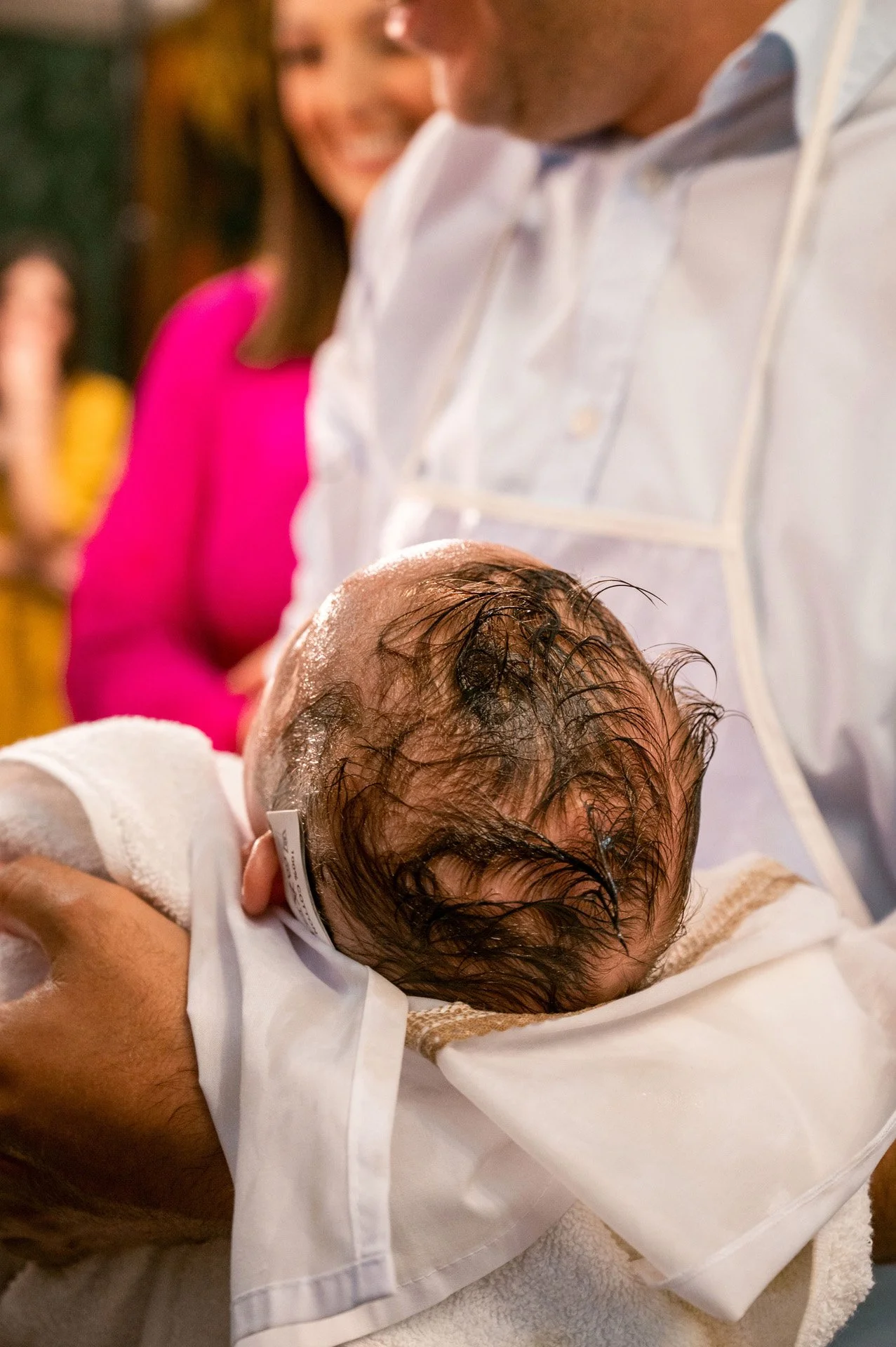 A newborn baby with wet hair being held by a person in a white shirt during a baptism or christening ceremony, with a woman in a pink top smiling in the background.