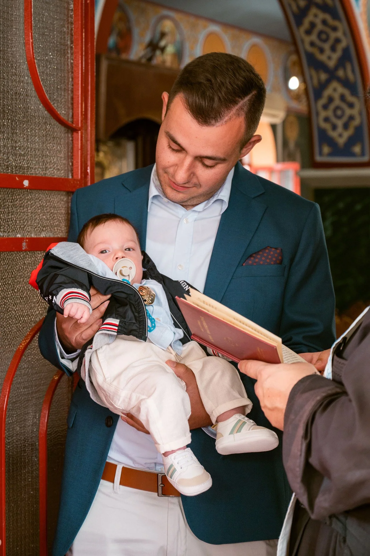 A man in a blue jacket holding a baby with a pacifier, while reading a document or book in a decorated indoor setting.
