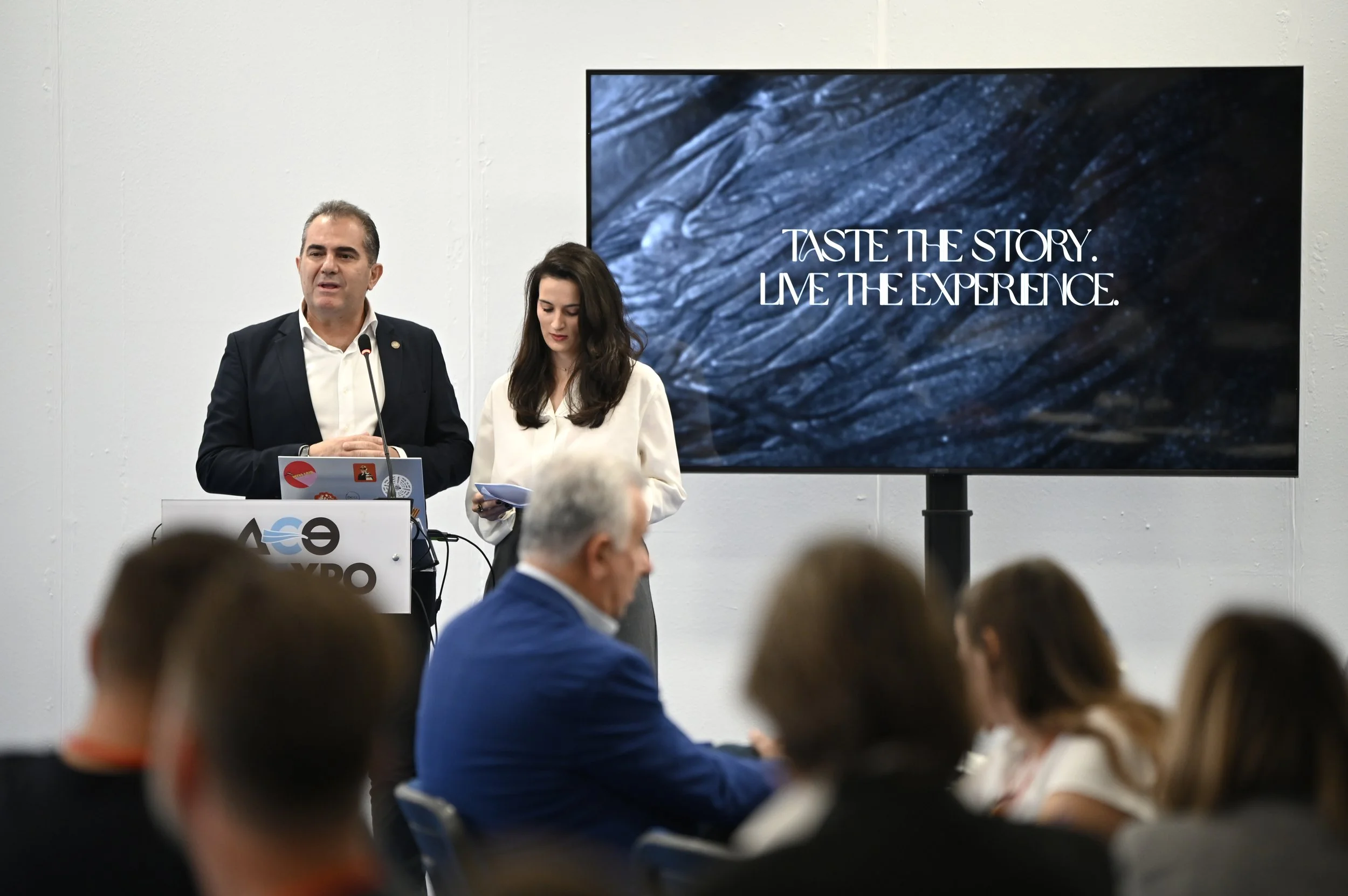 Two presenters at a conference, a man and a woman, standing in front of a large screen displaying the text "Taste the story. Live the experience." The man is speaking into a microphone, and the woman is looking at notes. There are audience members se