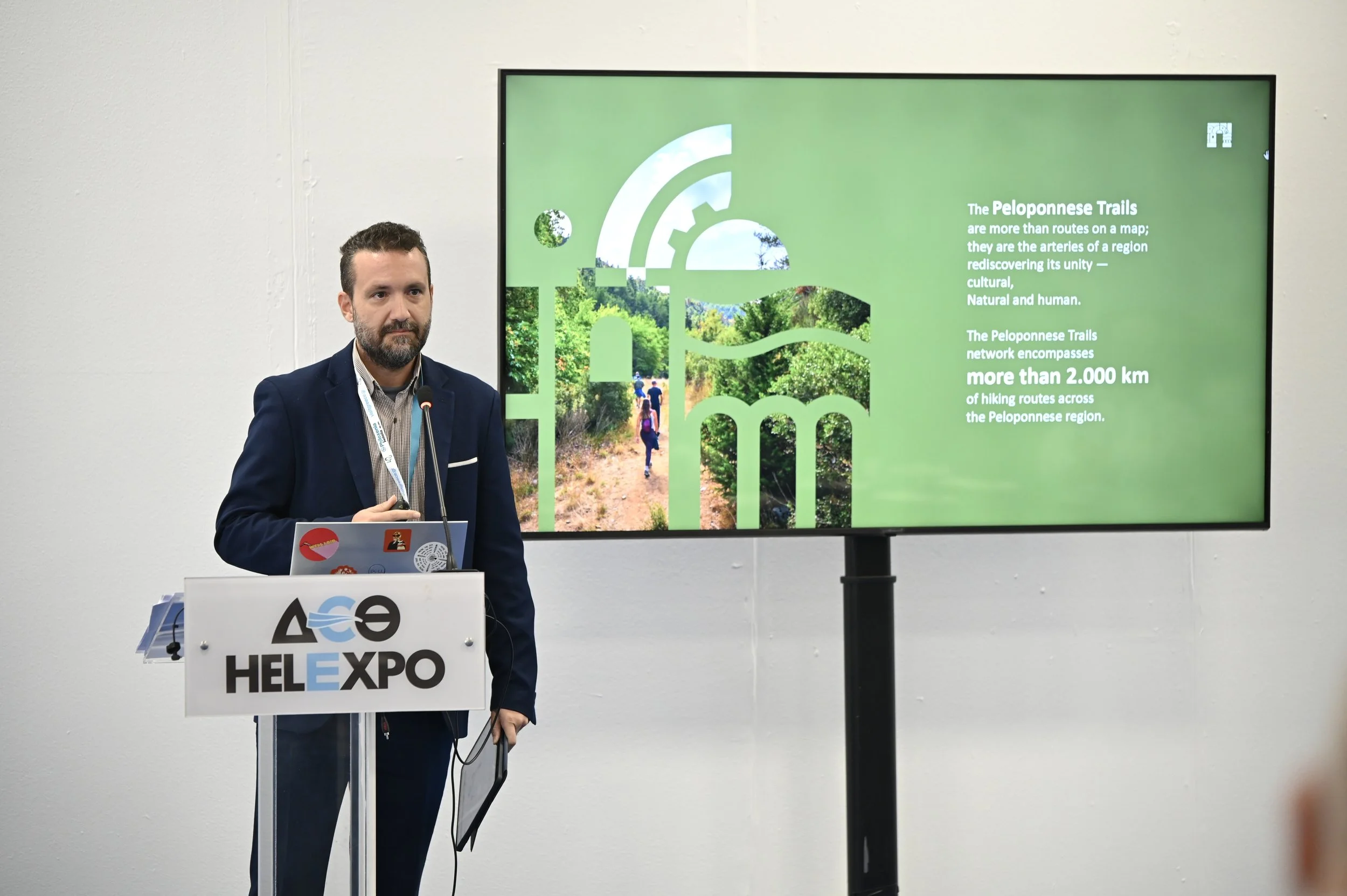 A man in a suit and tie standing at a podium with a microphone, giving a presentation about the Peloponnese Trails, with a large screen displaying information and images of hiking trails behind him.