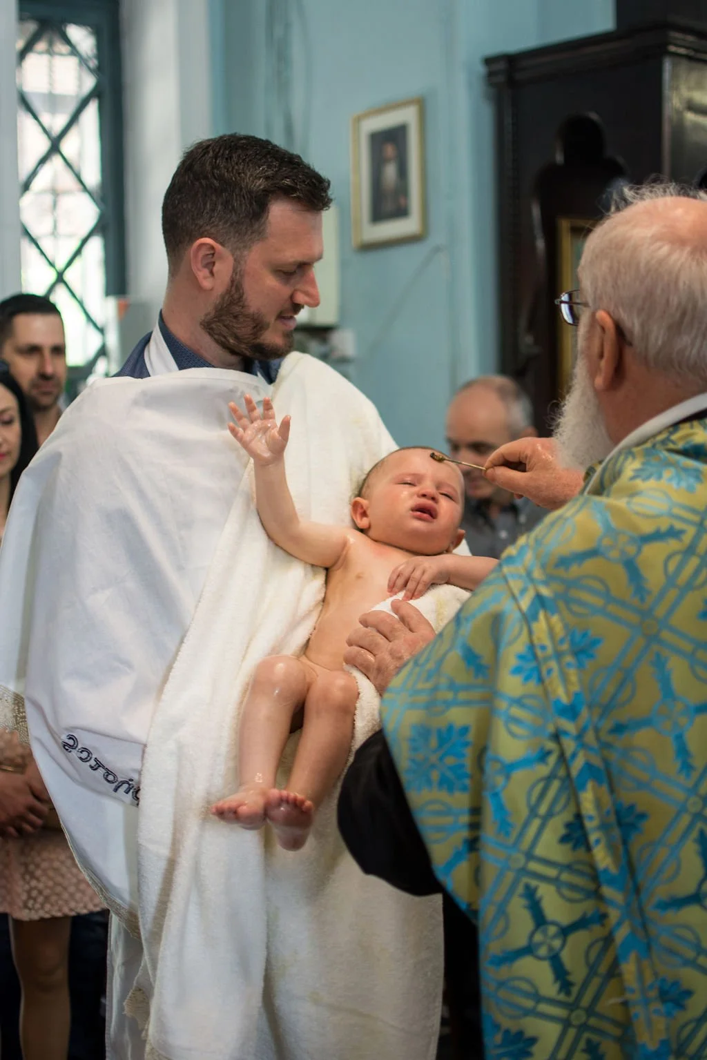 A baby is being baptized in a church, held by a man in a white cloth, while a priest in blue and yellow robes pours water on the baby’s head as others watch.