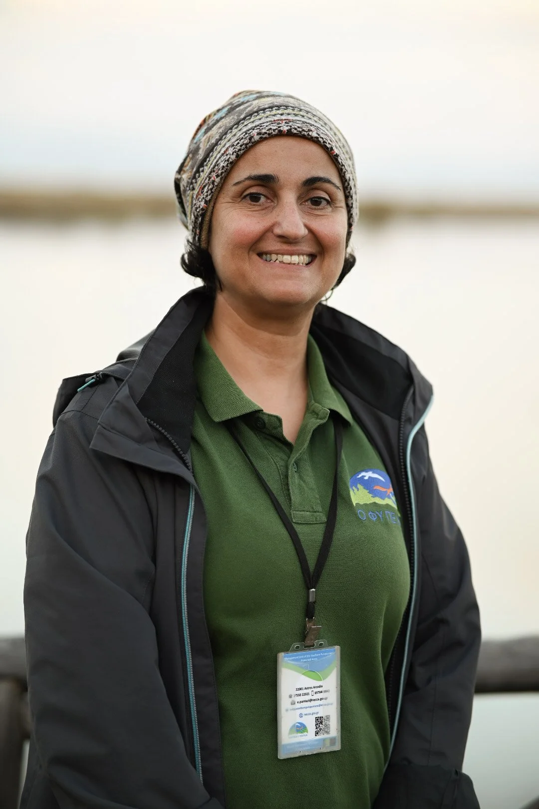 Woman wearing a patterned headscarf, green polo shirt with a logo, and a black jacket, standing outdoors near a body of water with a blurred natural background.