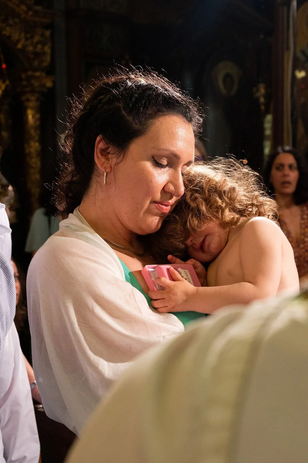 A woman with dark curly hair holding a young child with curly blond hair and no shirt, resting their head on her shoulder in a warm embrace inside a dimly lit, ornate room.