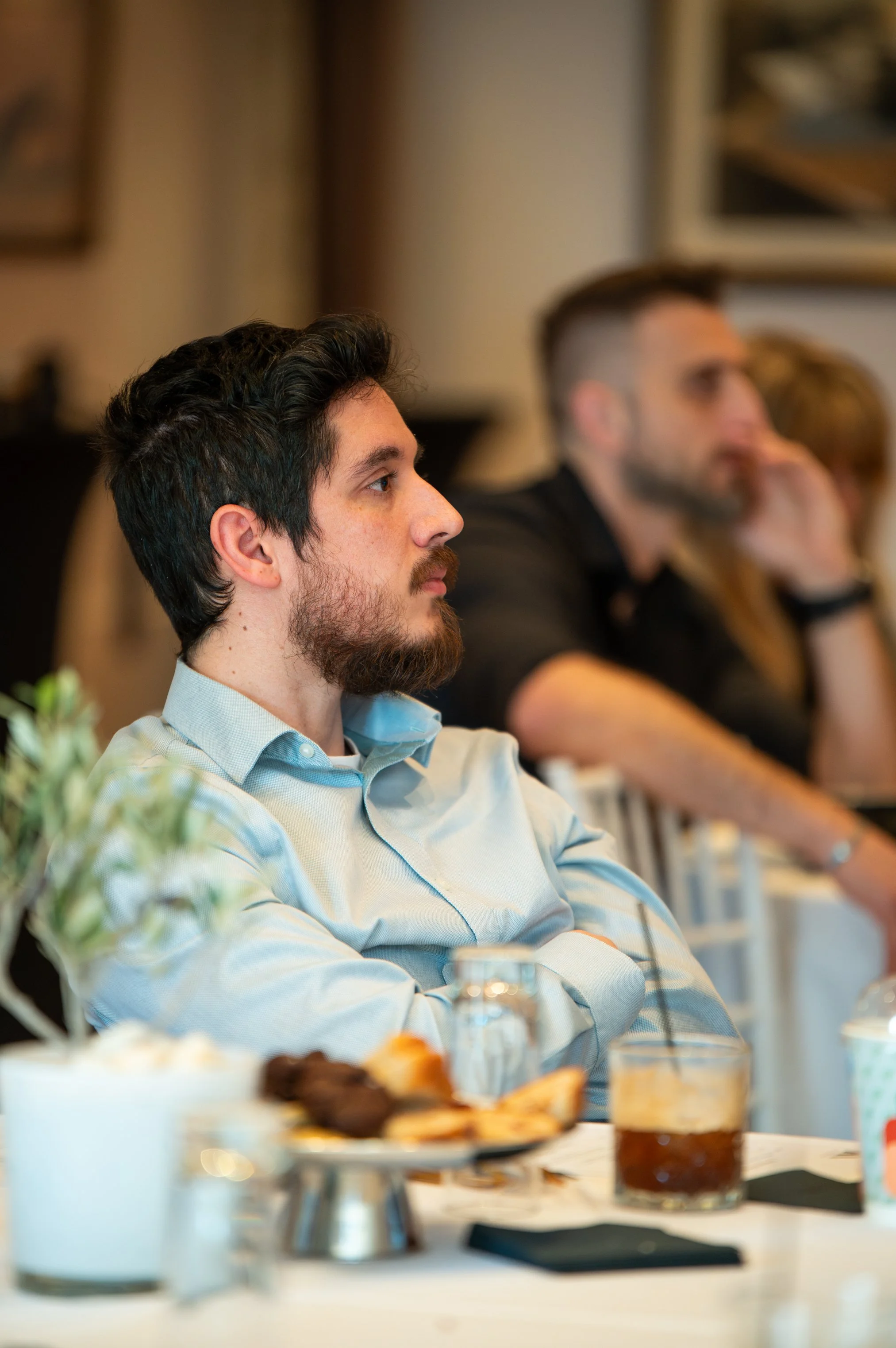 A man with dark hair and a beard, wearing a light blue shirt, is sitting at a table with food and drinks during a meeting or event. He is attentively listening, with other attendees visible in the background.