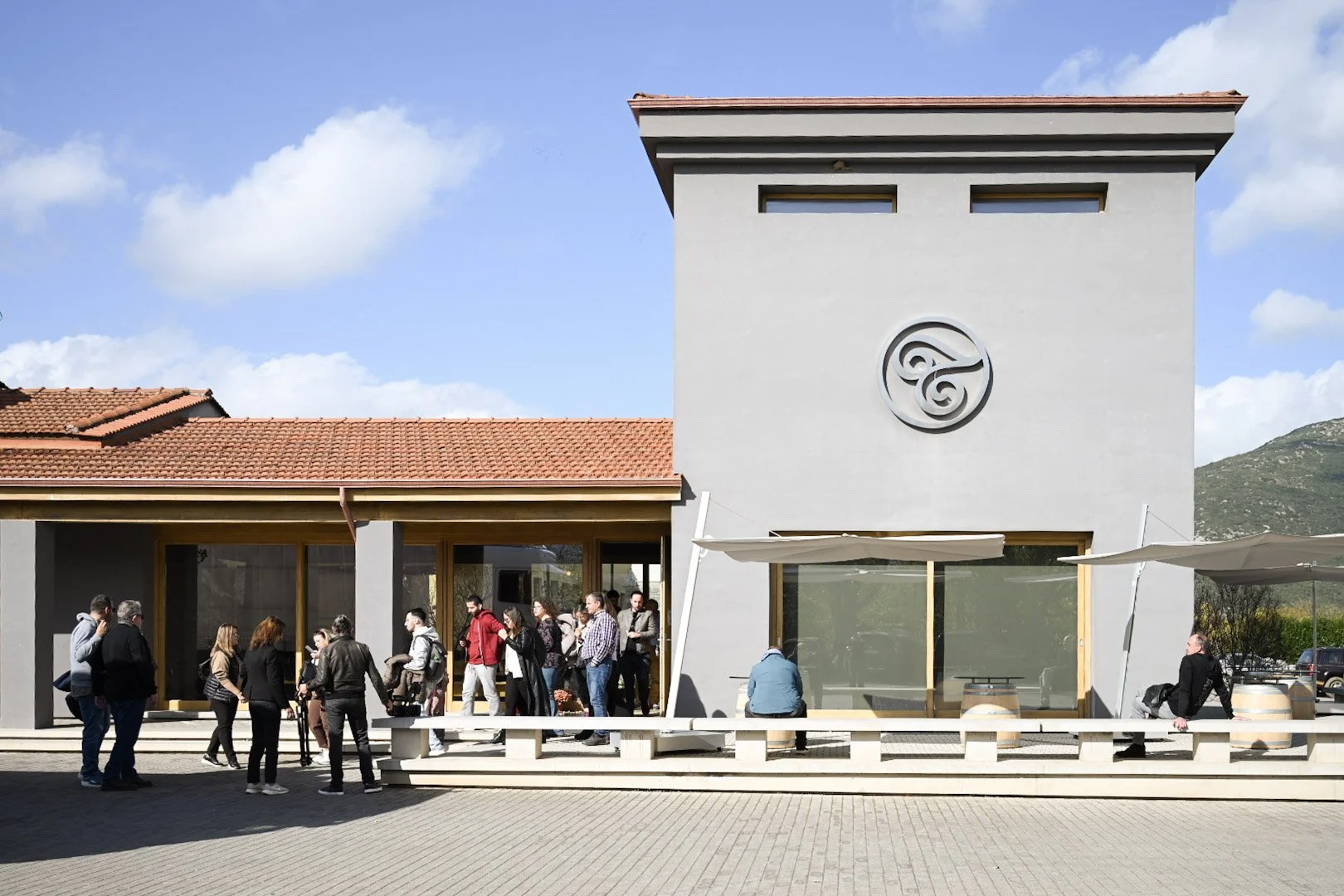 People standing and sitting outside a modern building with large windows and a circular logo on the wall, under a partly cloudy sky.