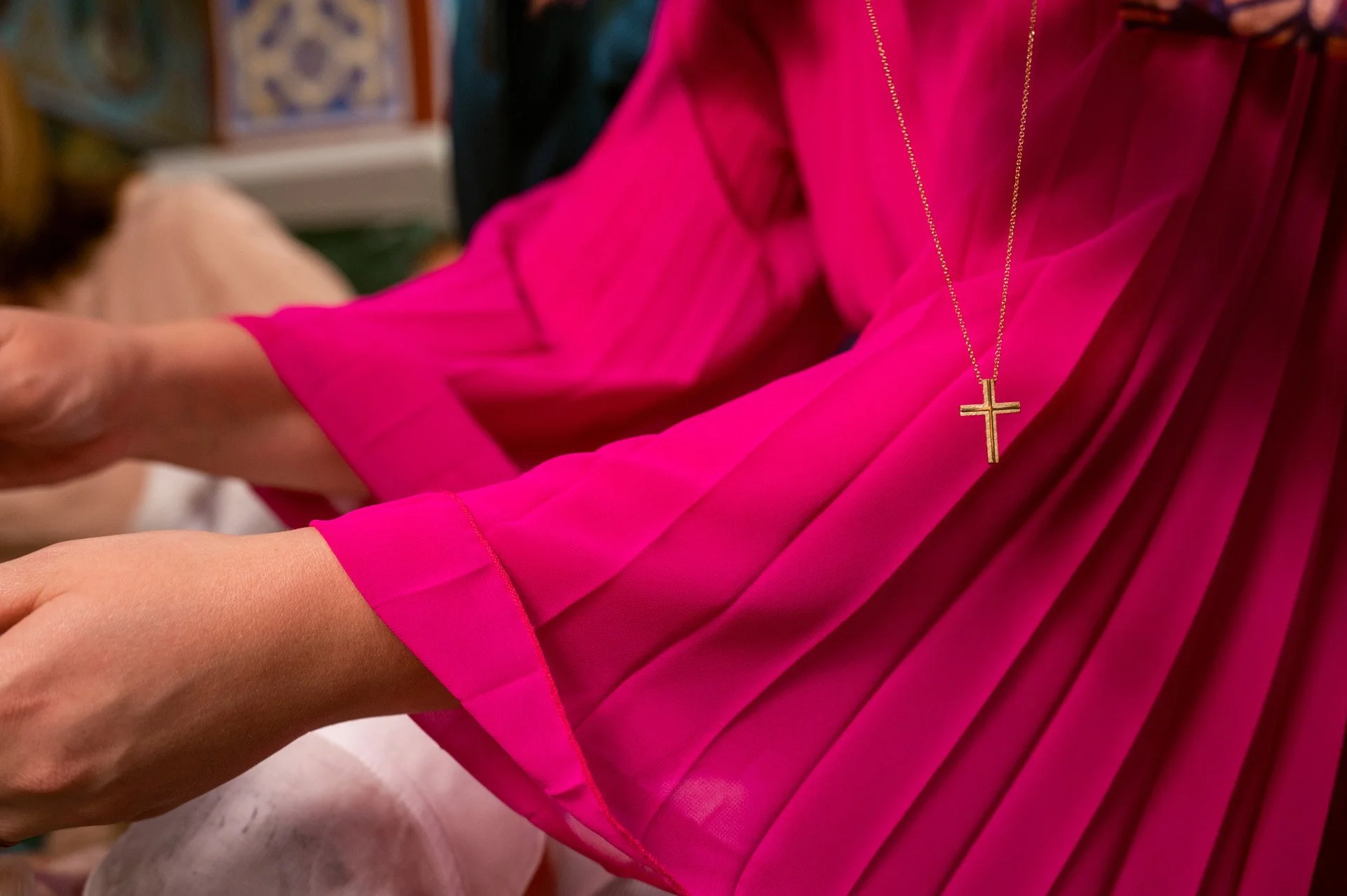 Close-up of a woman wearing a bright pink dress with pleated sleeves and a gold chain with a cross pendant around her neck. She appears to be participating in a religious or ceremonial activity.