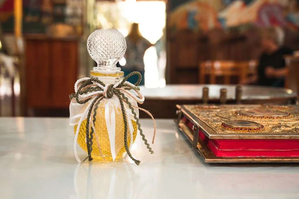 A decorative glass perfume bottle with a textured round stopper, wrapped in a cloth and tied with a ribbon, placed on a white table next to an ornate, colorful book.