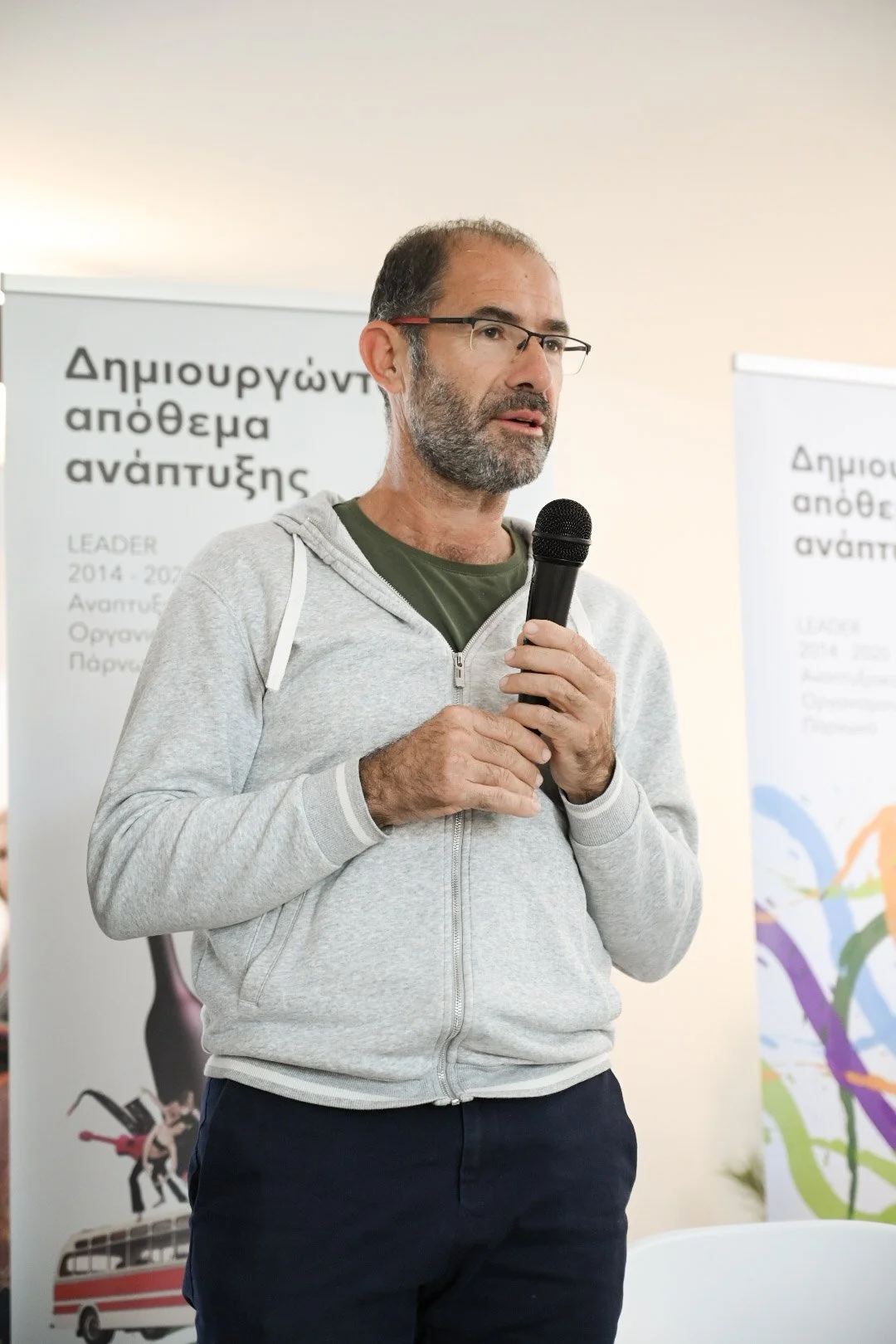 A man with glasses, beard, and grey hair, wearing a grey hoodie over a green shirt, holding a microphone and speaking at an indoor event, with banners in the background containing Greek text.