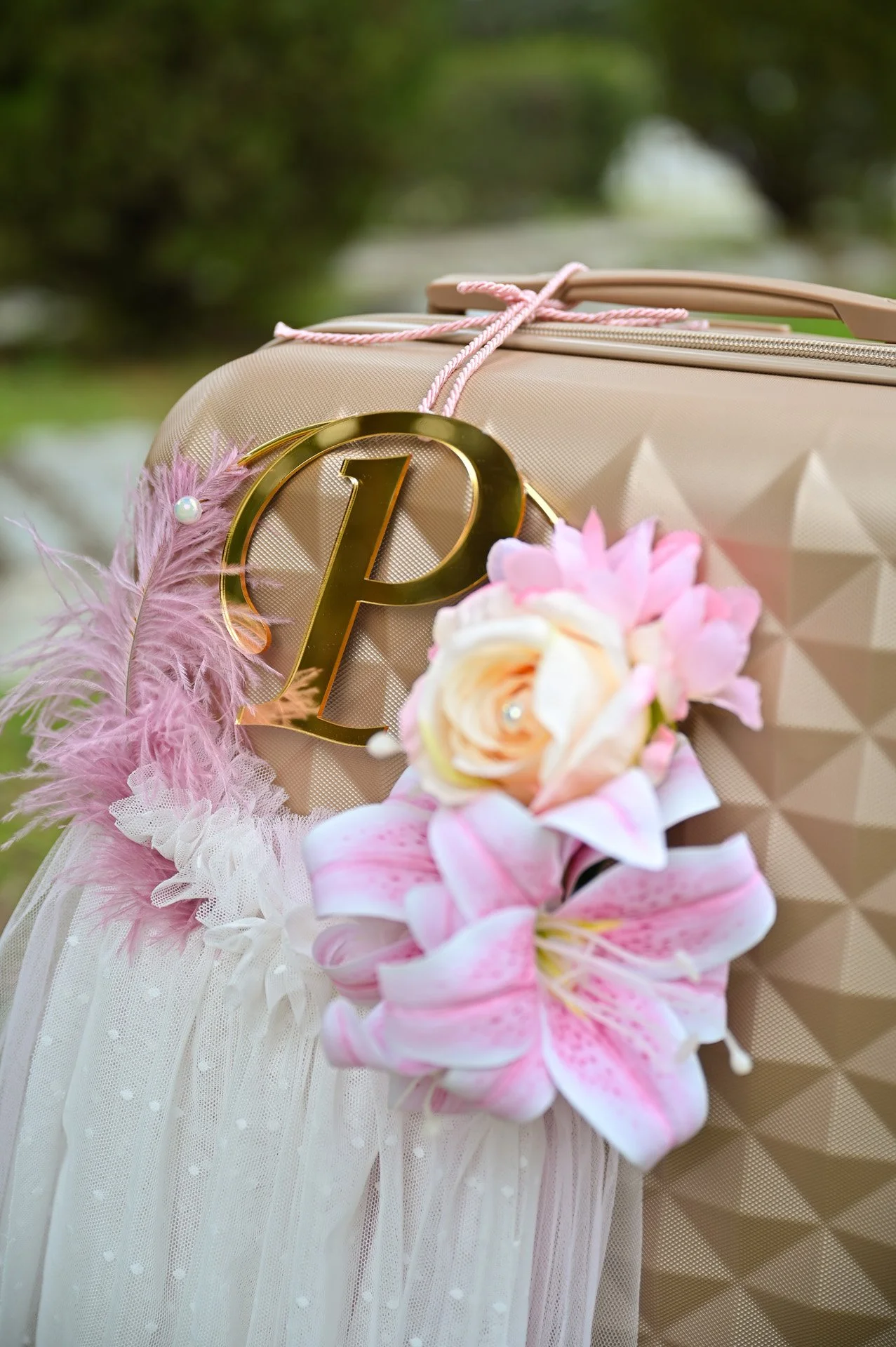 A beige suitcase decorated with pink feathers, a pink flower, and a gold monogrammed initial 'P' for a wedding or special event.