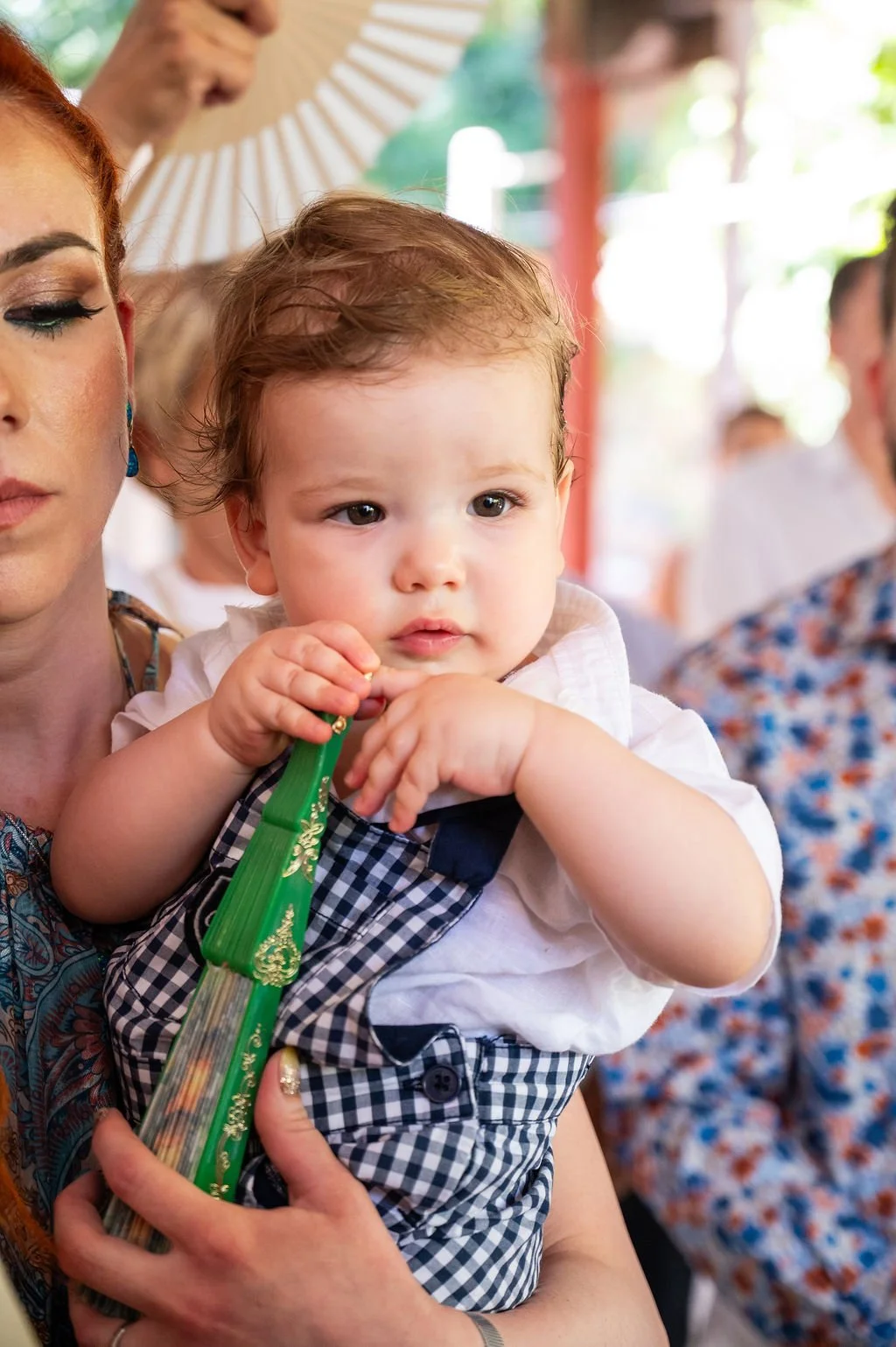 A young child with light brown hair, dressed in a white shirt and checkered overalls, held by a woman with red hair at an outdoor gathering, with a casual and colorful background.
