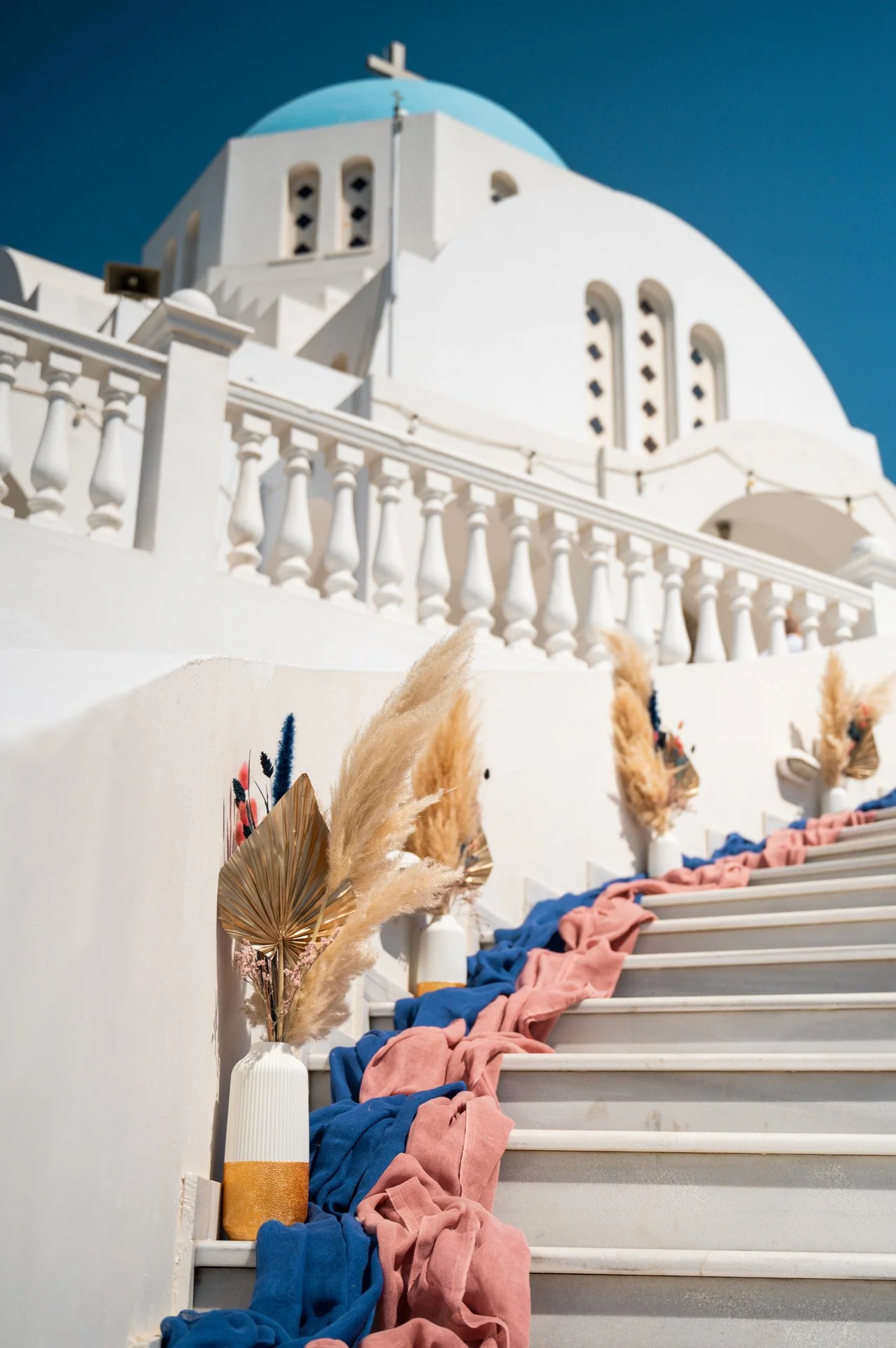 Decorated staircase with vases holding pampas grass and draped fabric leading up to a white Greek-style building with a blue dome.