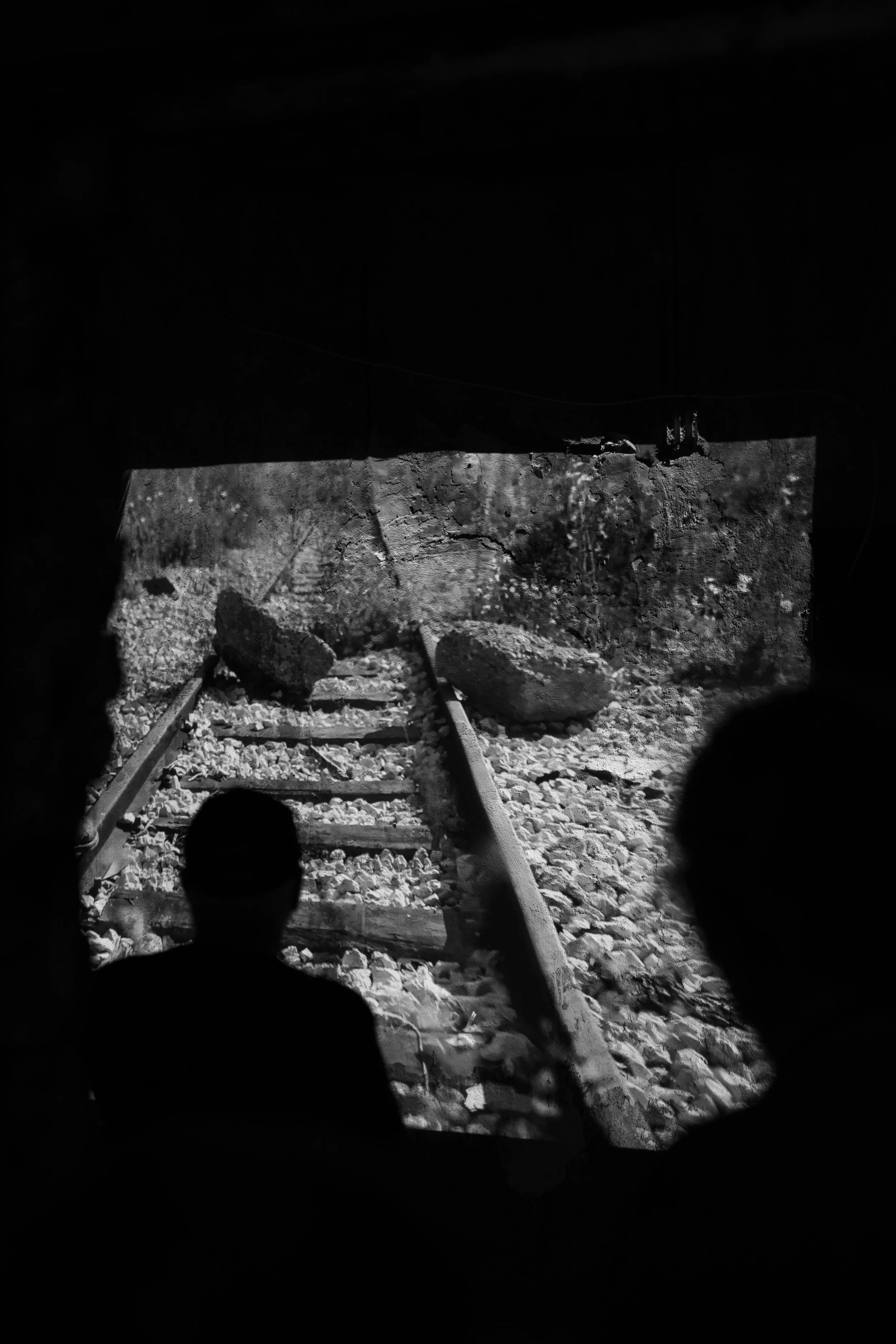 Two silhouetted people observing a black-and-white projected image of damaged train tracks with rocks and large boulders.