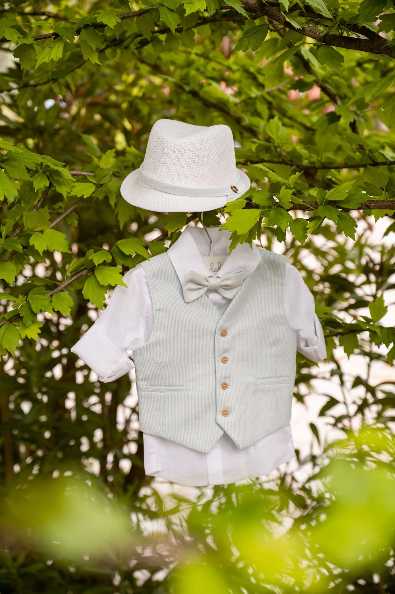A child's white dress shirt, light gray vest, matching bow tie, and white fedora hat hanging on a hanger among green leaves.