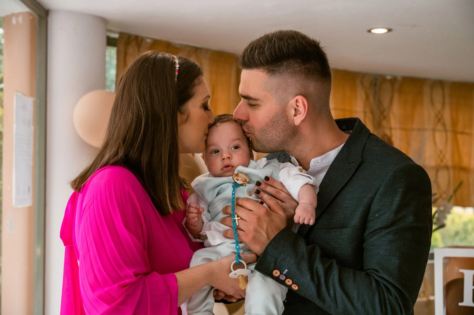 A family of three with a woman, a man, and a baby, sharing a moment of affection indoors. Both adults are leaning in and kissing the baby's head.