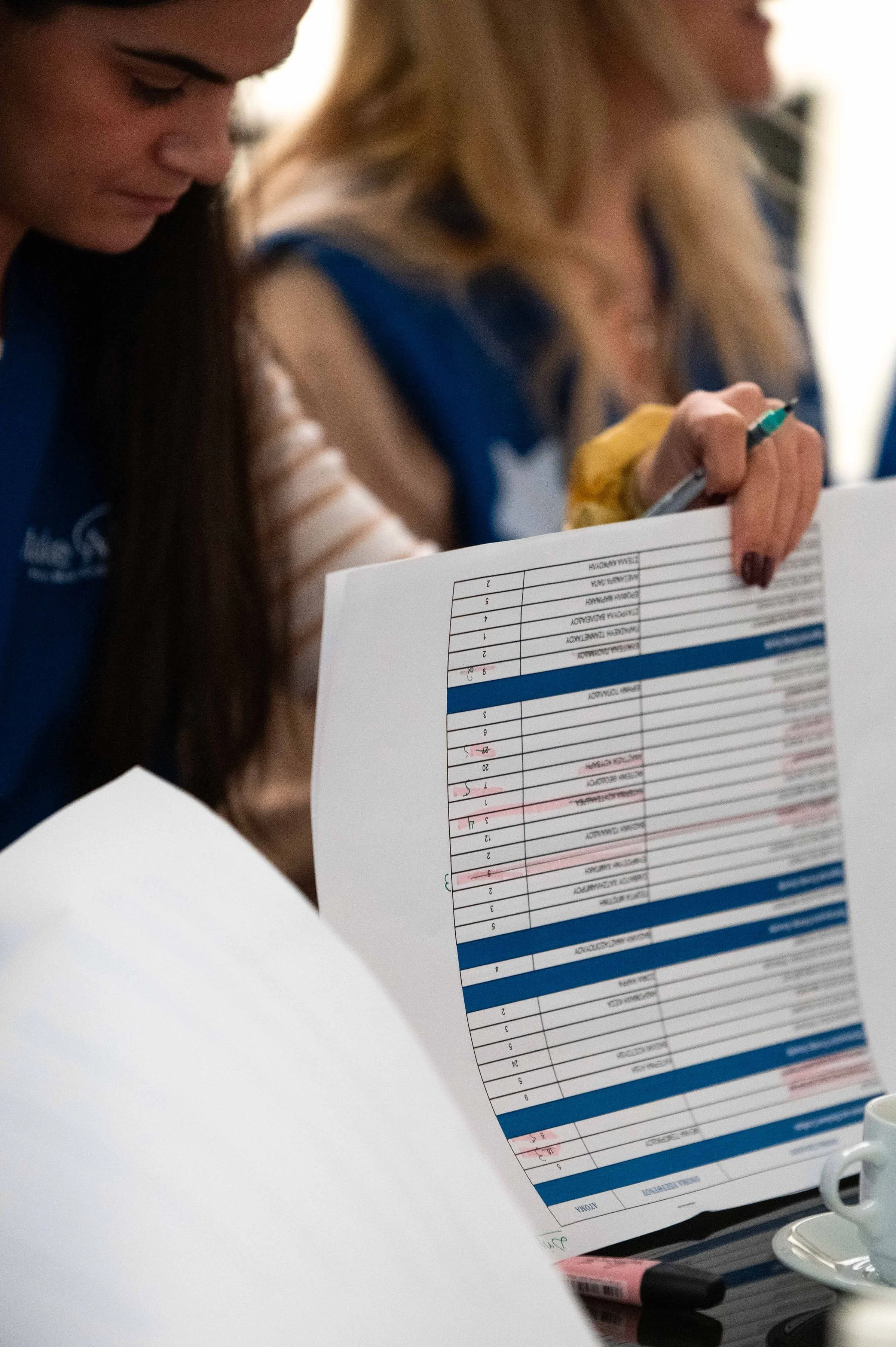 Two women reviewing a printed schedule or document, one holding a pen, in an indoor setting.