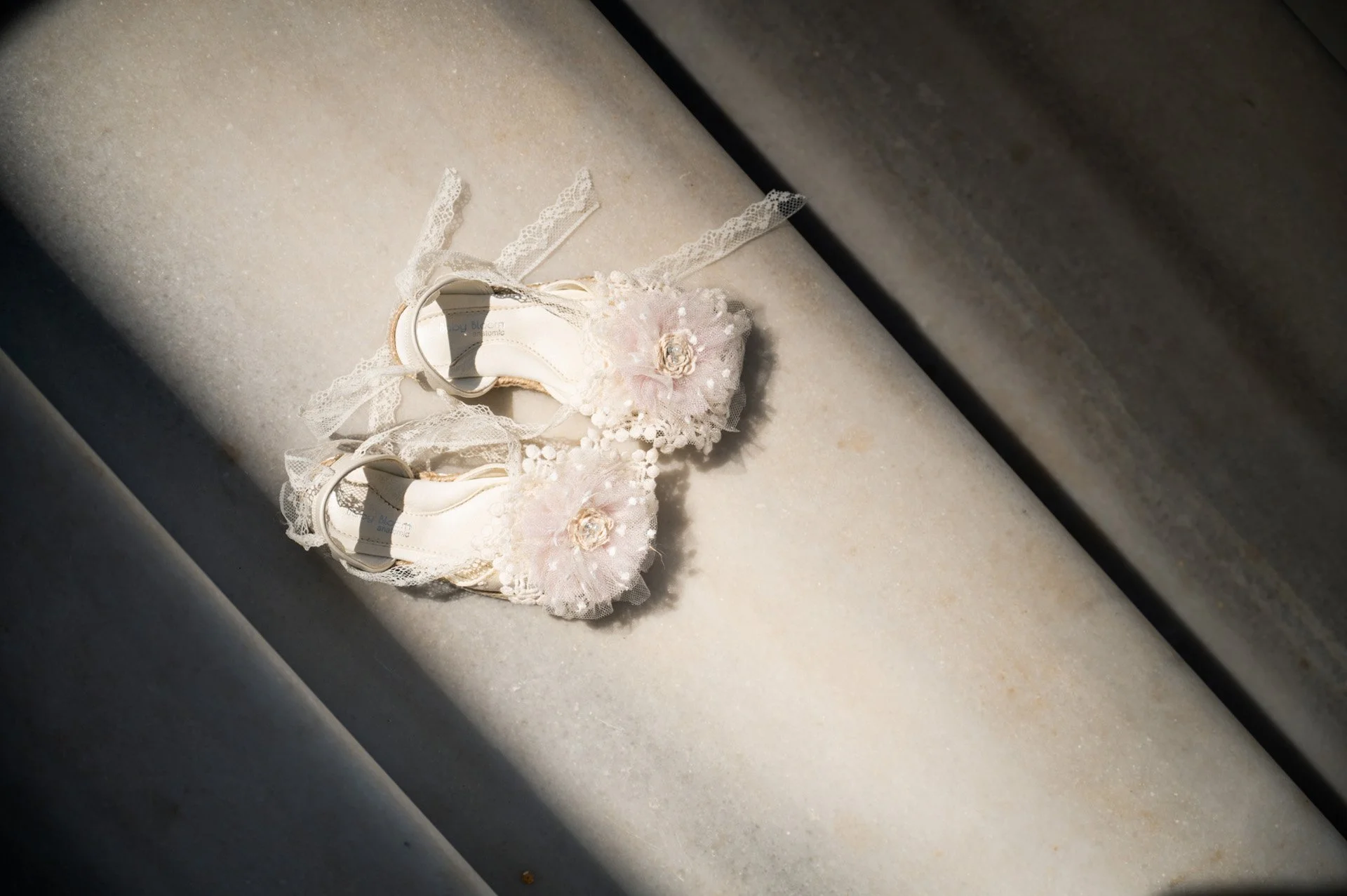 Pair of white bridal shoes with lace and floral embellishments placed on a marble surface.