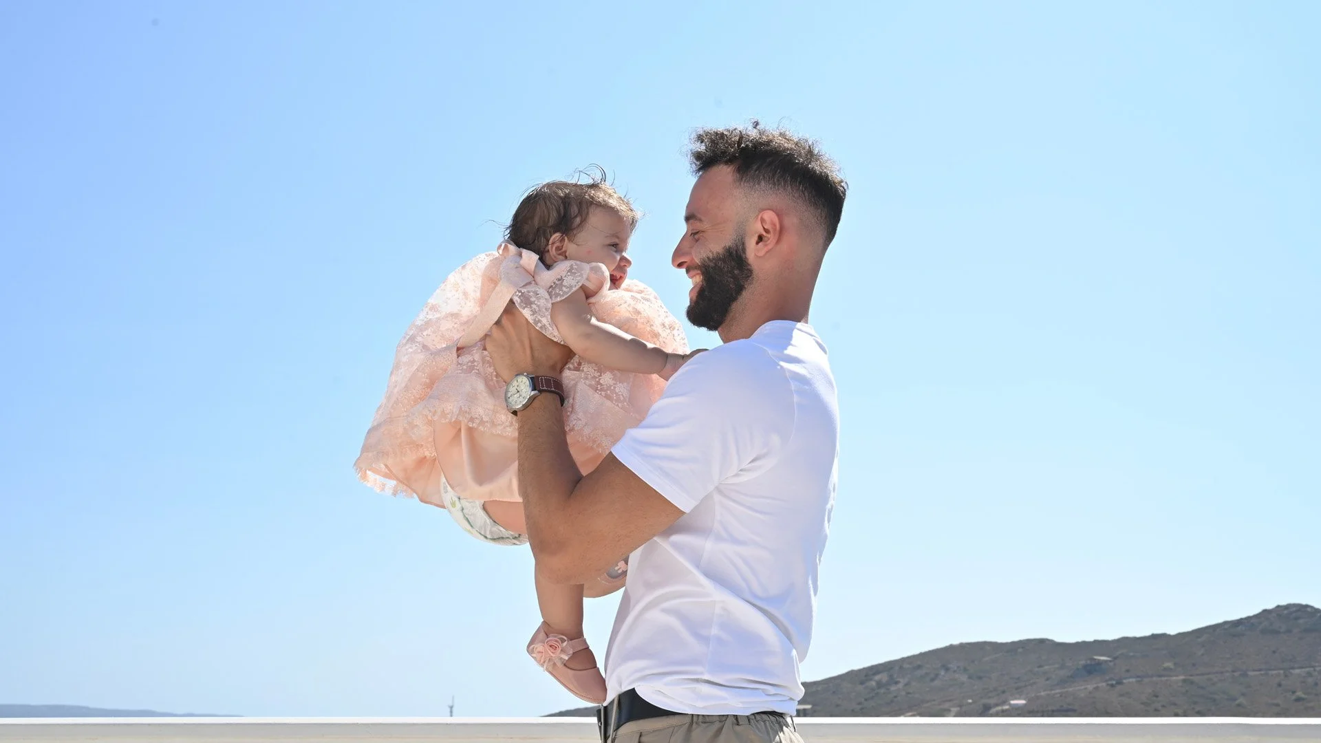 A man holding a young girl in a pink dress, both smiling and looking at each other, against a clear blue sky and distant hills.