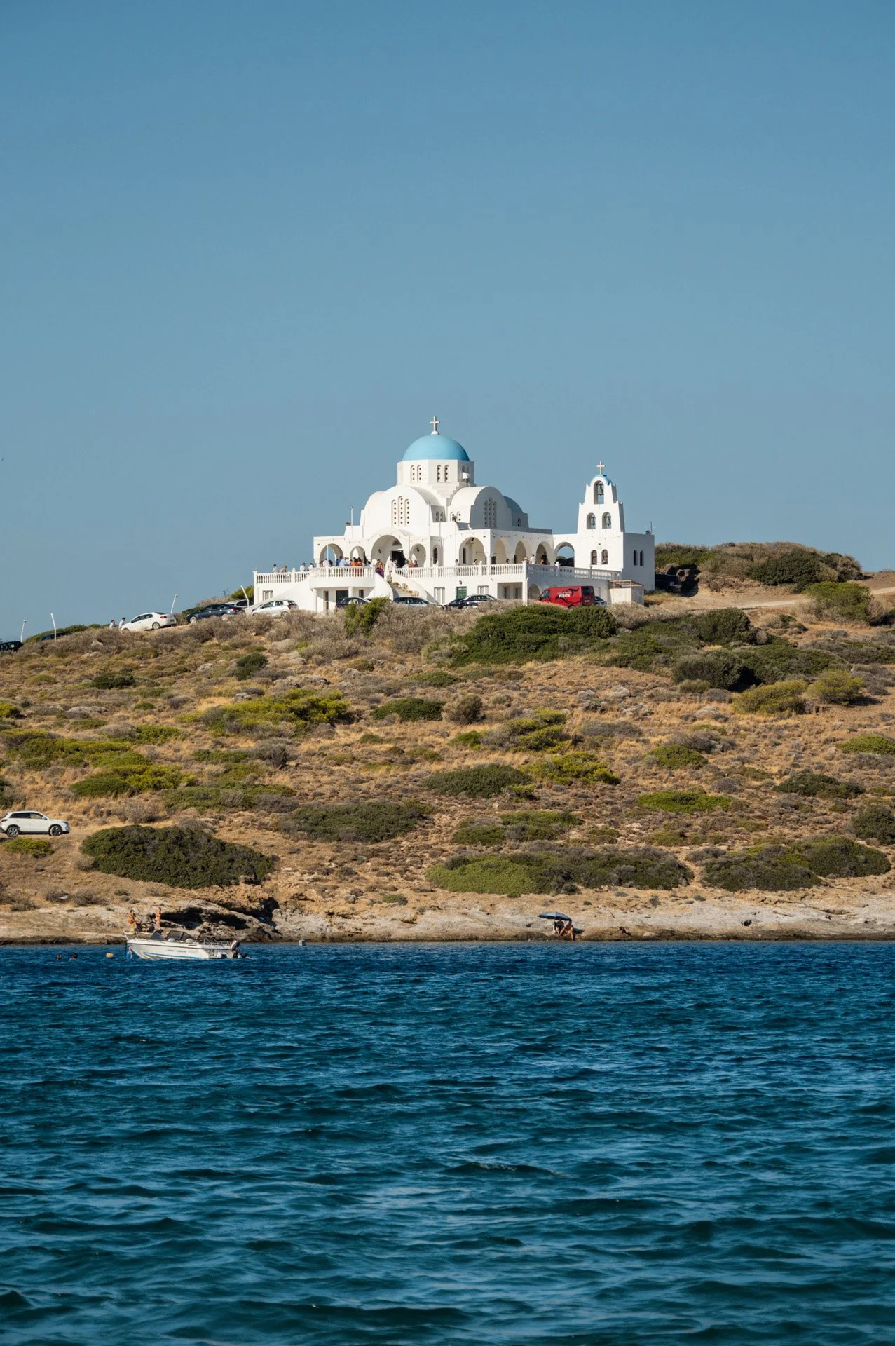 A white church with a blue dome and several crosses on top, situated on a hillside overlooking a body of water, with a few cars parked nearby and a boat on the water.