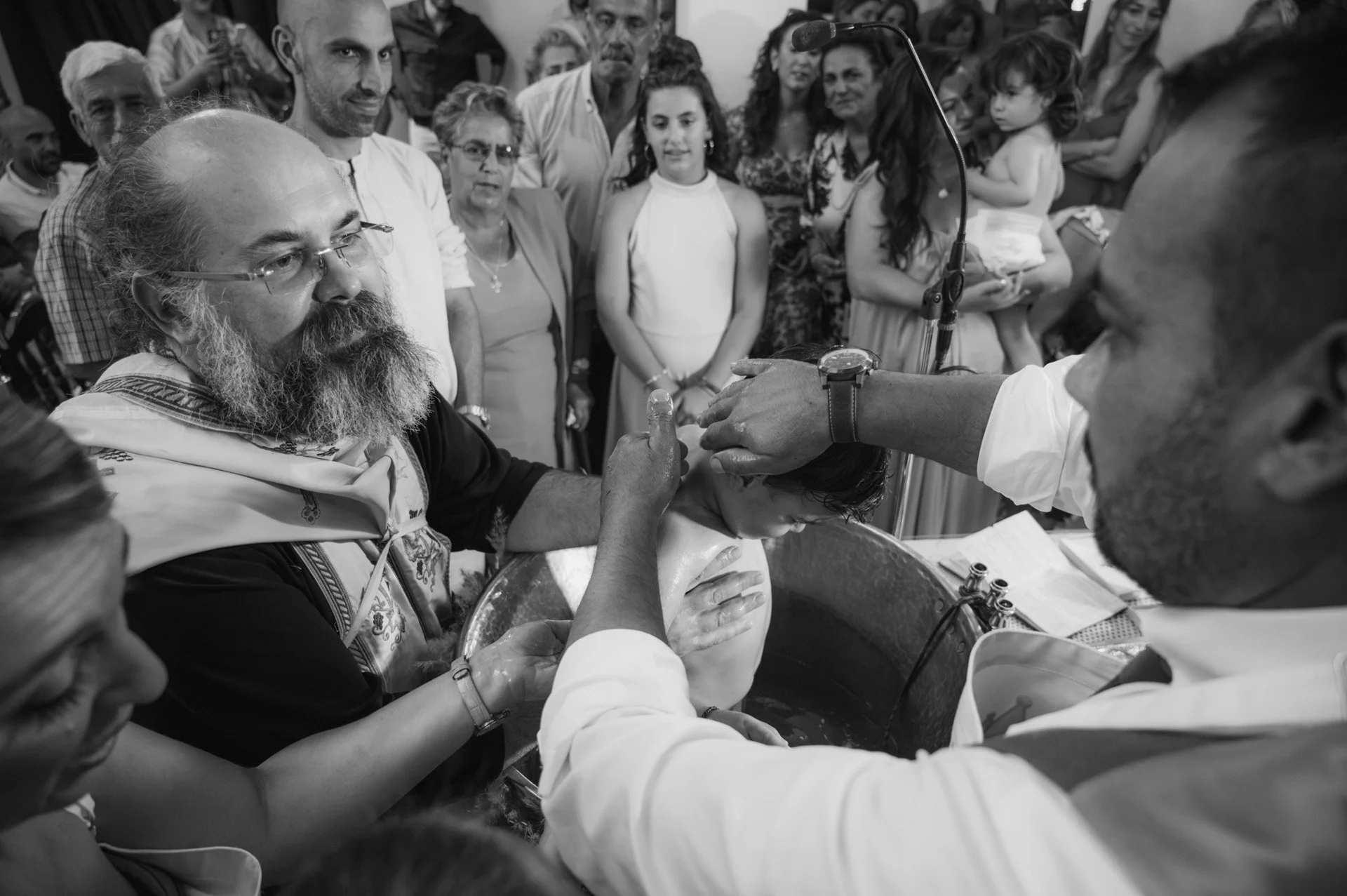 A religious baptism ceremony with a person immersed in water while a priest and others witness around a baptismal pool.