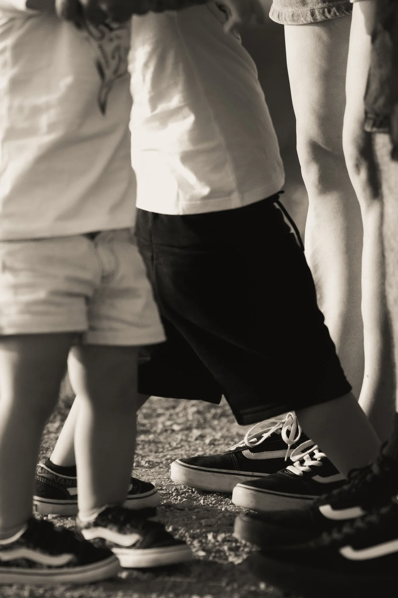 Close-up of children wearing casual shorts and sneakers, standing outdoors on a gravel surface.