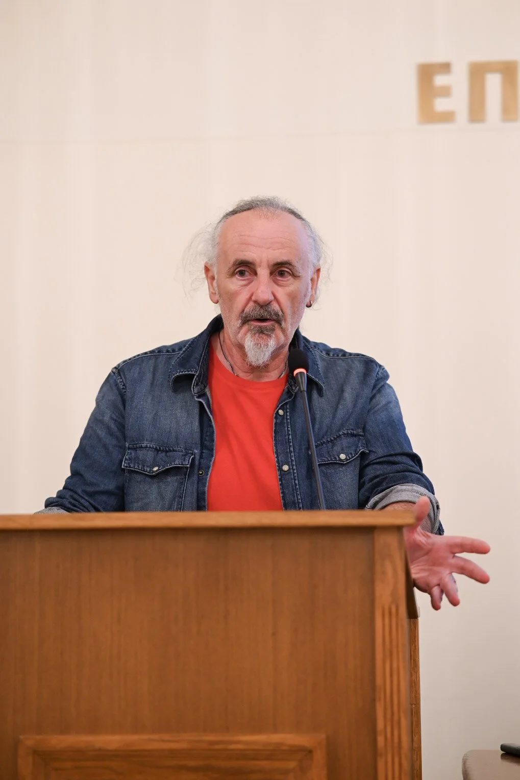 A man with gray hair and beard wearing a red shirt and denim jacket speaking behind a podium with a microphone.