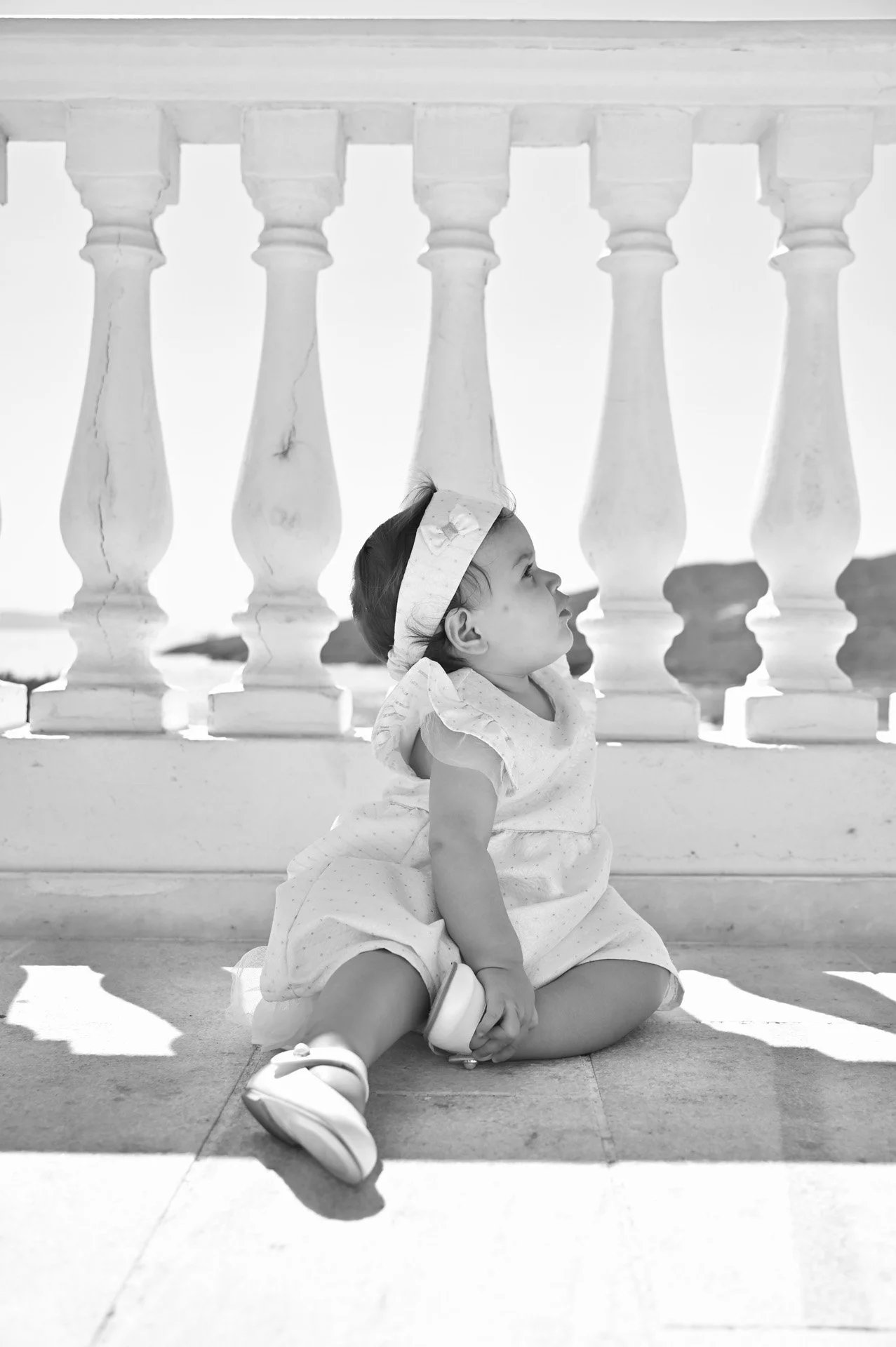 A young girl in a dress with a headband sitting on the ground, looking up at a stone railing with balusters.