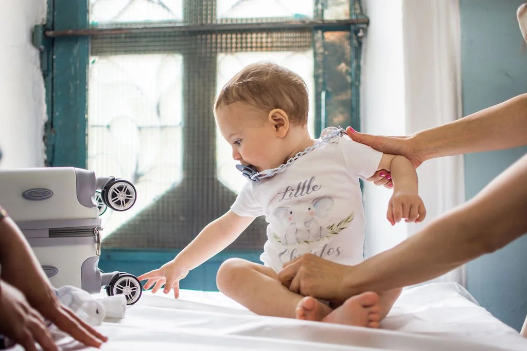 A young child with light brown hair, wearing a white t-shirt with an elephant and the words "Little Elefantes," is sitting on a bed as an adult helps or supports them. An adult's hand is gently touching the child's shoulder. There is a white suitcase