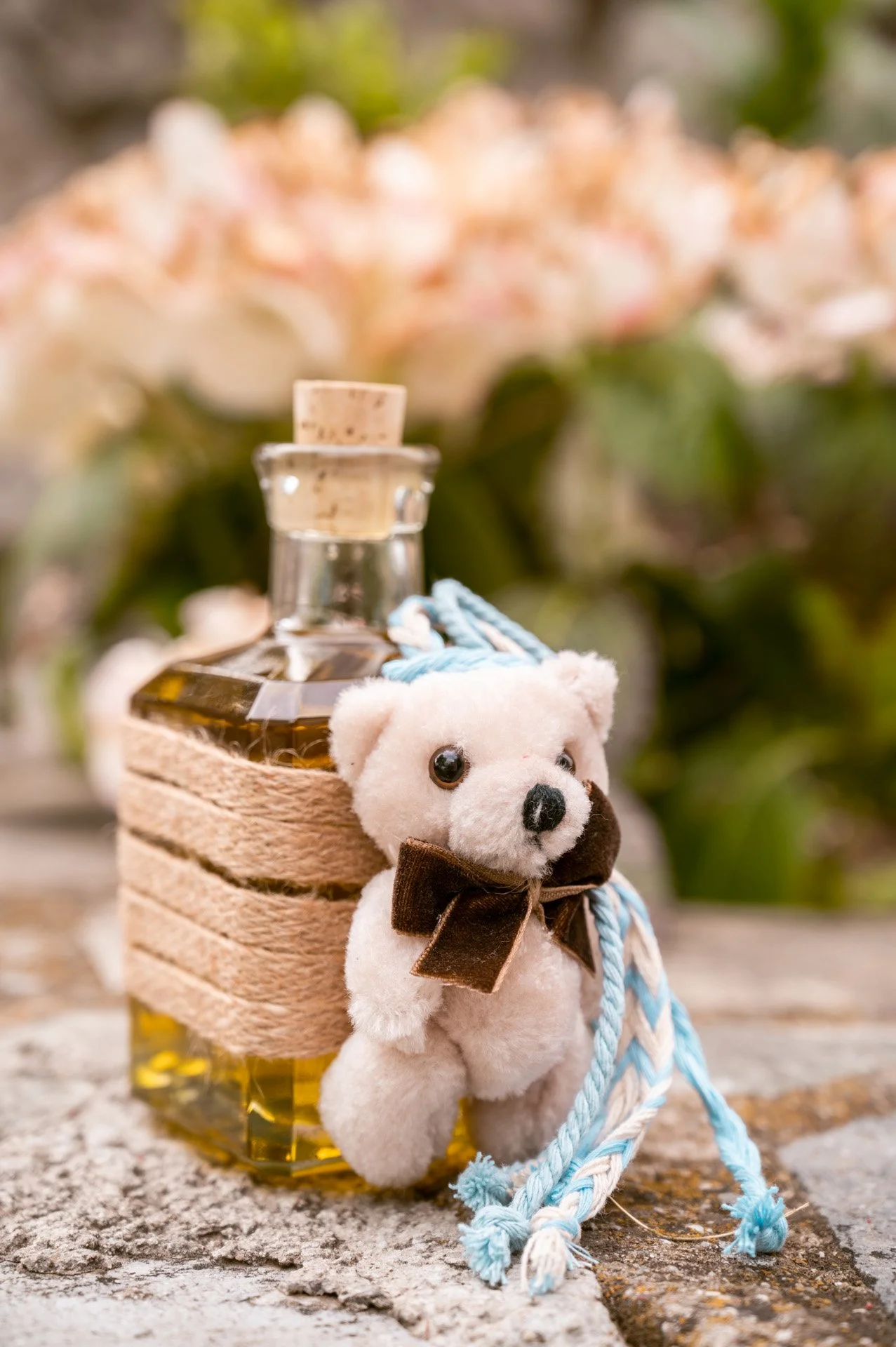 A small plush teddy bear with a brown bow tie attached to a bottle of oil, decorated with a cozy knit bag and hanging blue and white braided cords, placed on a rustic surface with a blurred garden background.