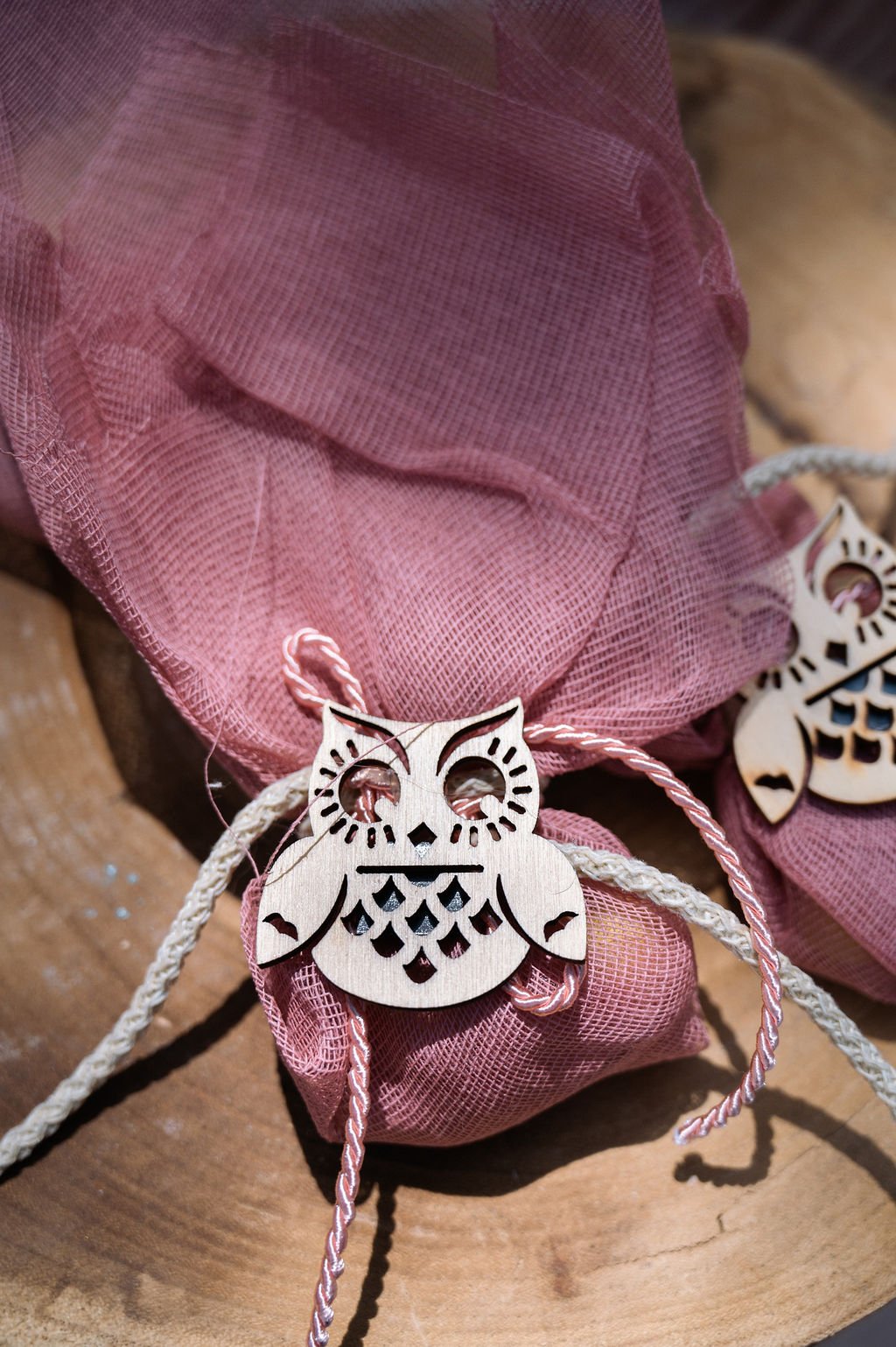 Wooden owl-shaped ornaments with cutout details on pink fabric pouches with drawstrings, placed on a wooden surface.