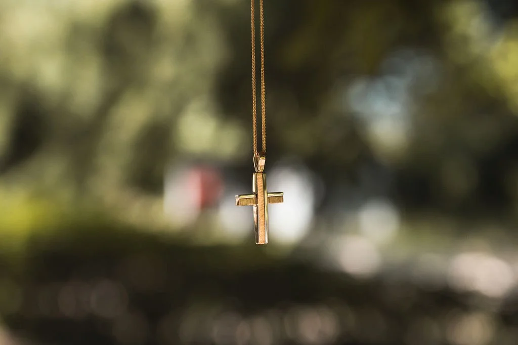 A gold cross necklace hanging outdoors against a blurred background of trees and sunlight.