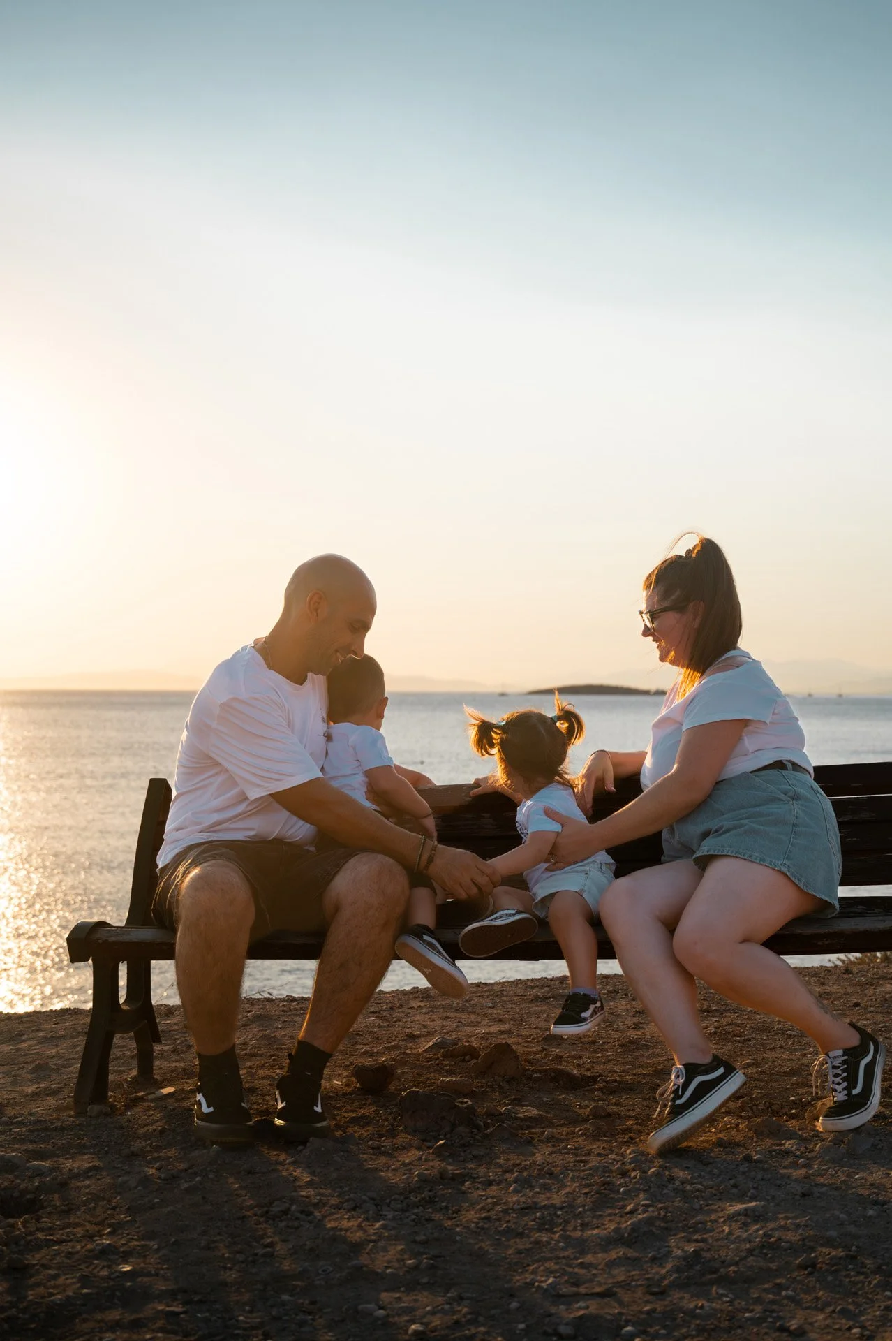 A family of four, including two young children, sitting on a bench by a body of water during sunset, playing and smiling together.