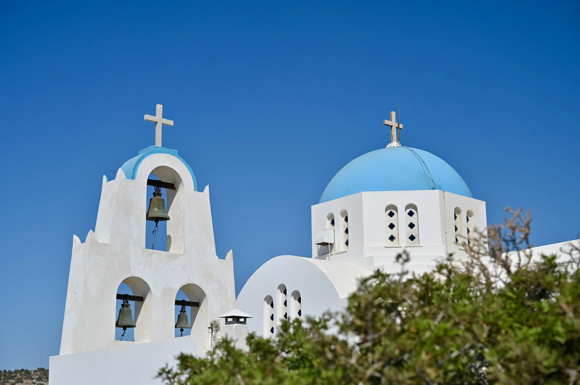 White church with a blue dome and bell tower in Santorini, Greece, against a clear blue sky.