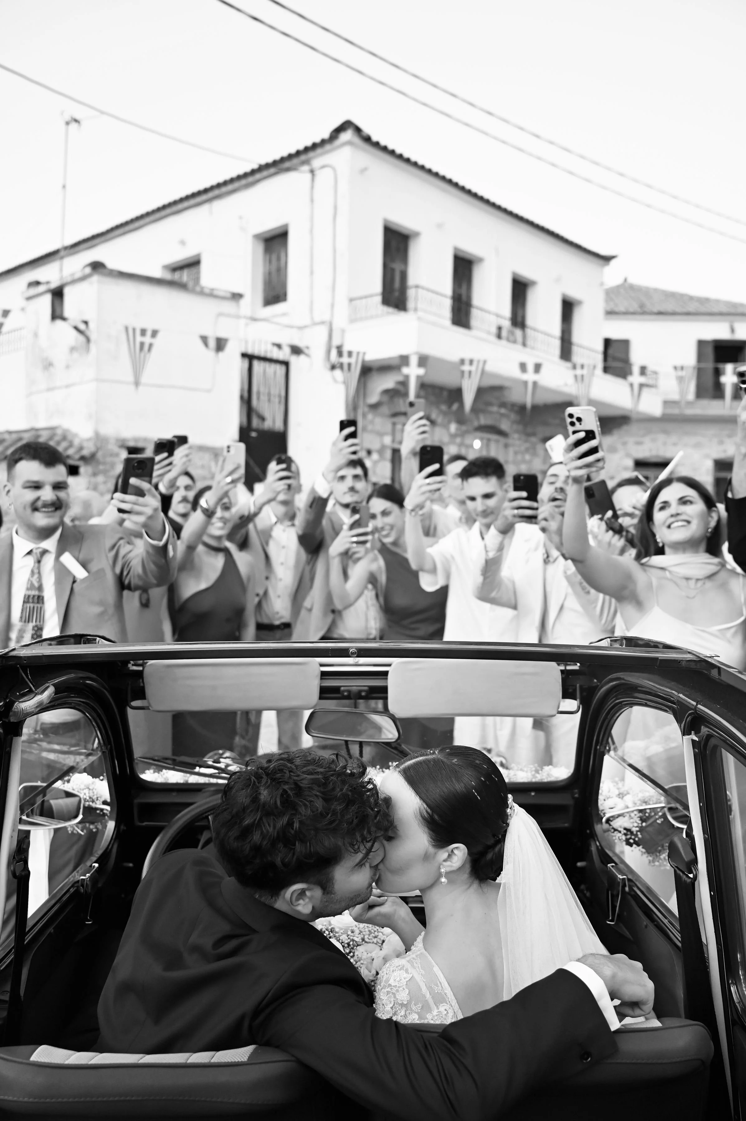 A couple kissing in a small car during their wedding with a crowd of people taking photos and videos in the background.