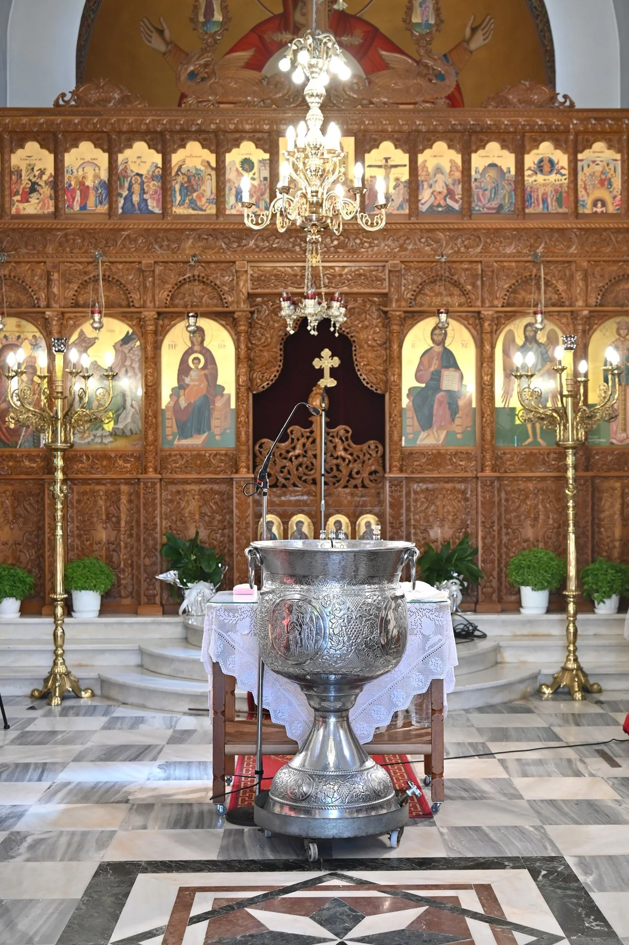 A church interior featuring an ornate wooden altar with religious icons, gold chandeliers, white potted plants, and a metal baptismal font on a stand, with marble flooring.