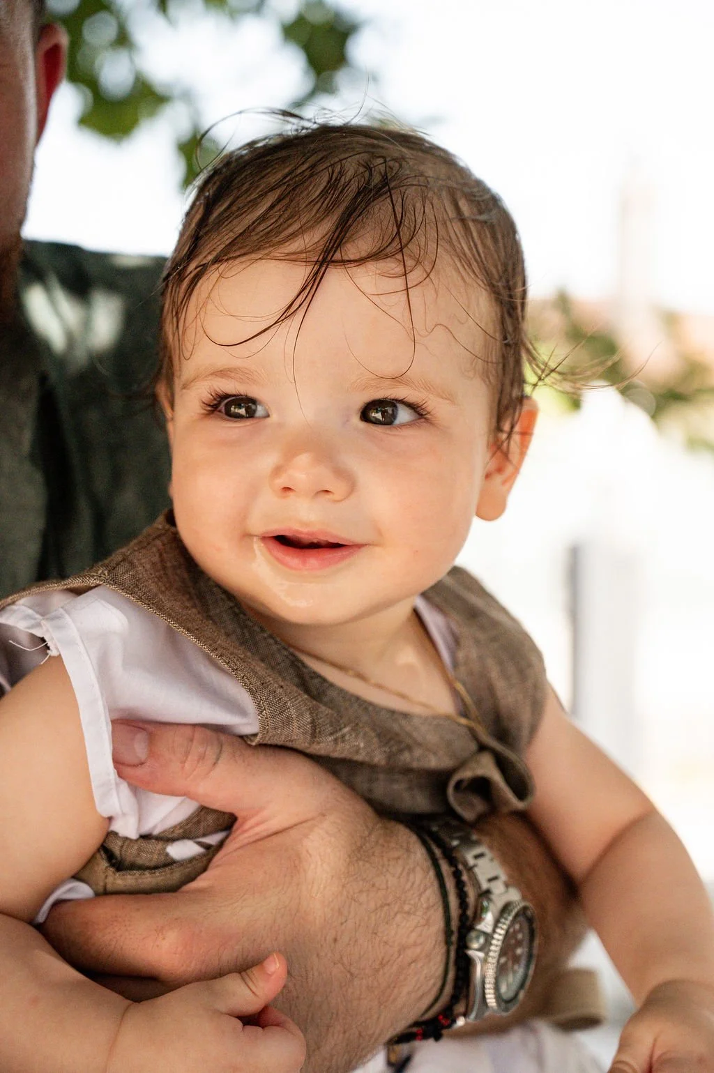 Close-up of a smiling baby with wet hair being held by an adult, outdoors, blurred trees in the background.