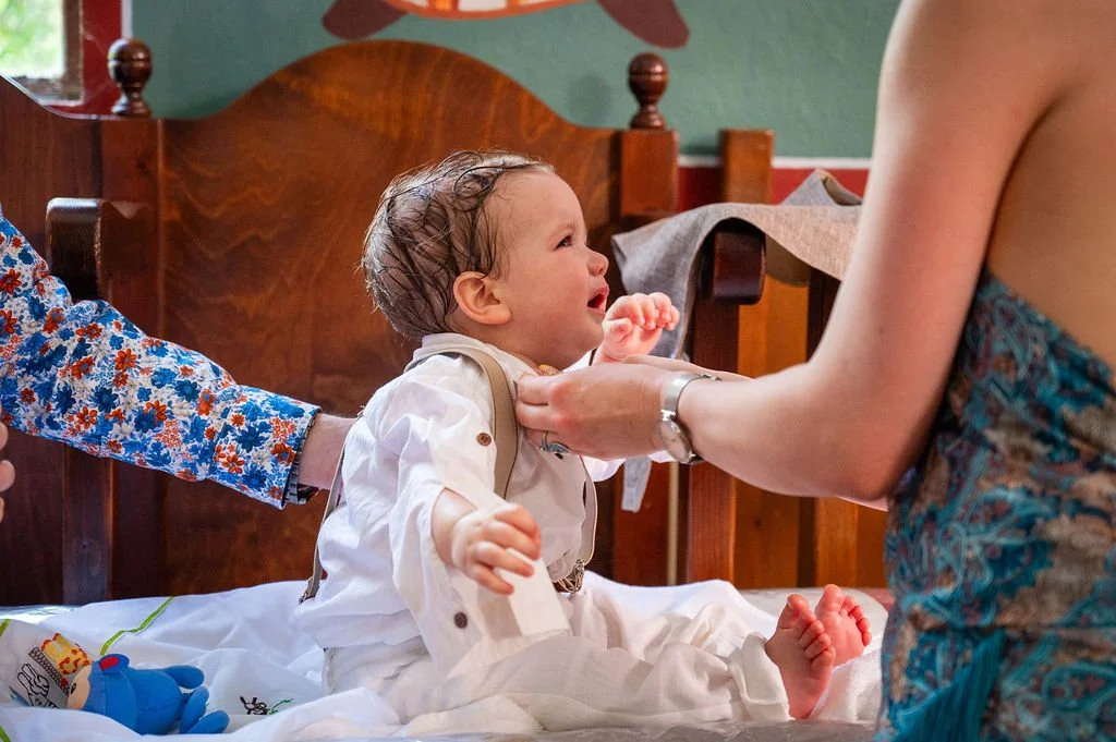 A young child with wet hair sitting on a bed, crying, while a woman in a strapless dress and a watch holds the child's chest, and another person wearing a floral shirt reaches out to hold the child's arm in a bedroom with wooden furniture.