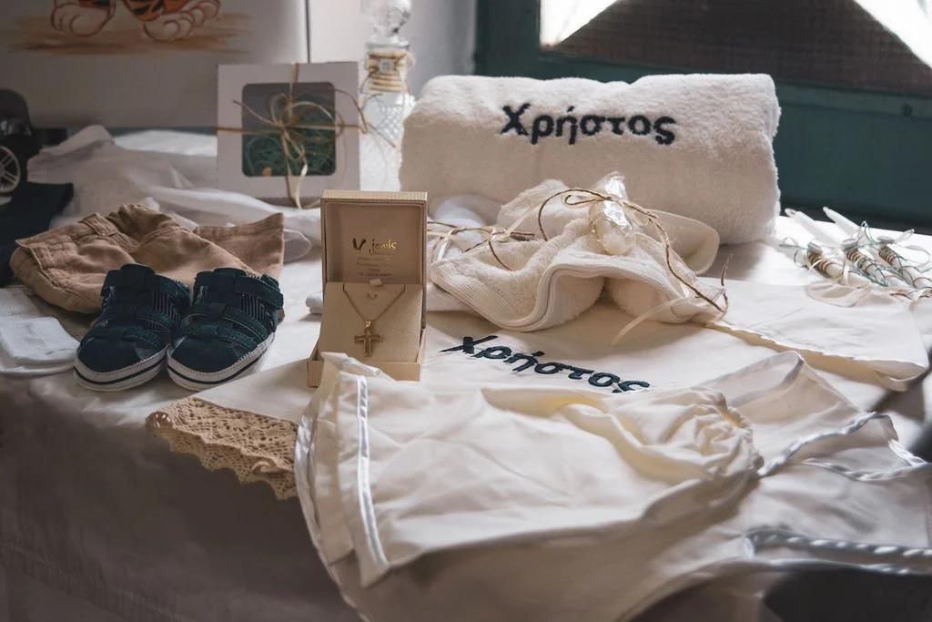 Baby clothes and accessories arranged on a table, including a white onesie with Greek script, a pair of tiny sneakers, a gold cross necklace, a white towel with Greek writing, and wrapped gifts.