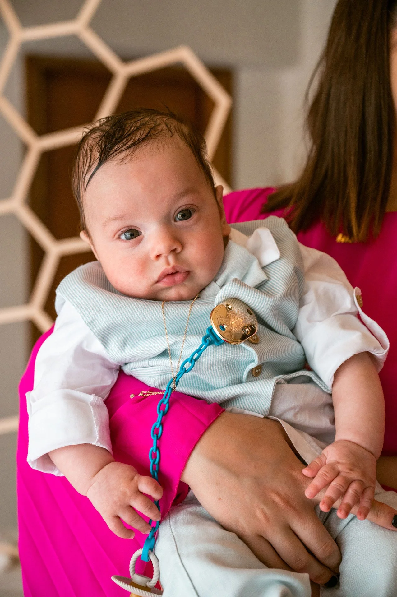 A baby with dark hair and big eyes, dressed in a white and light blue outfit, is sitting on an adult's lap. The baby is wearing a gold pacifier attached to a blue chain and is holding onto it with one hand. The adult is wearing a bright pink top.