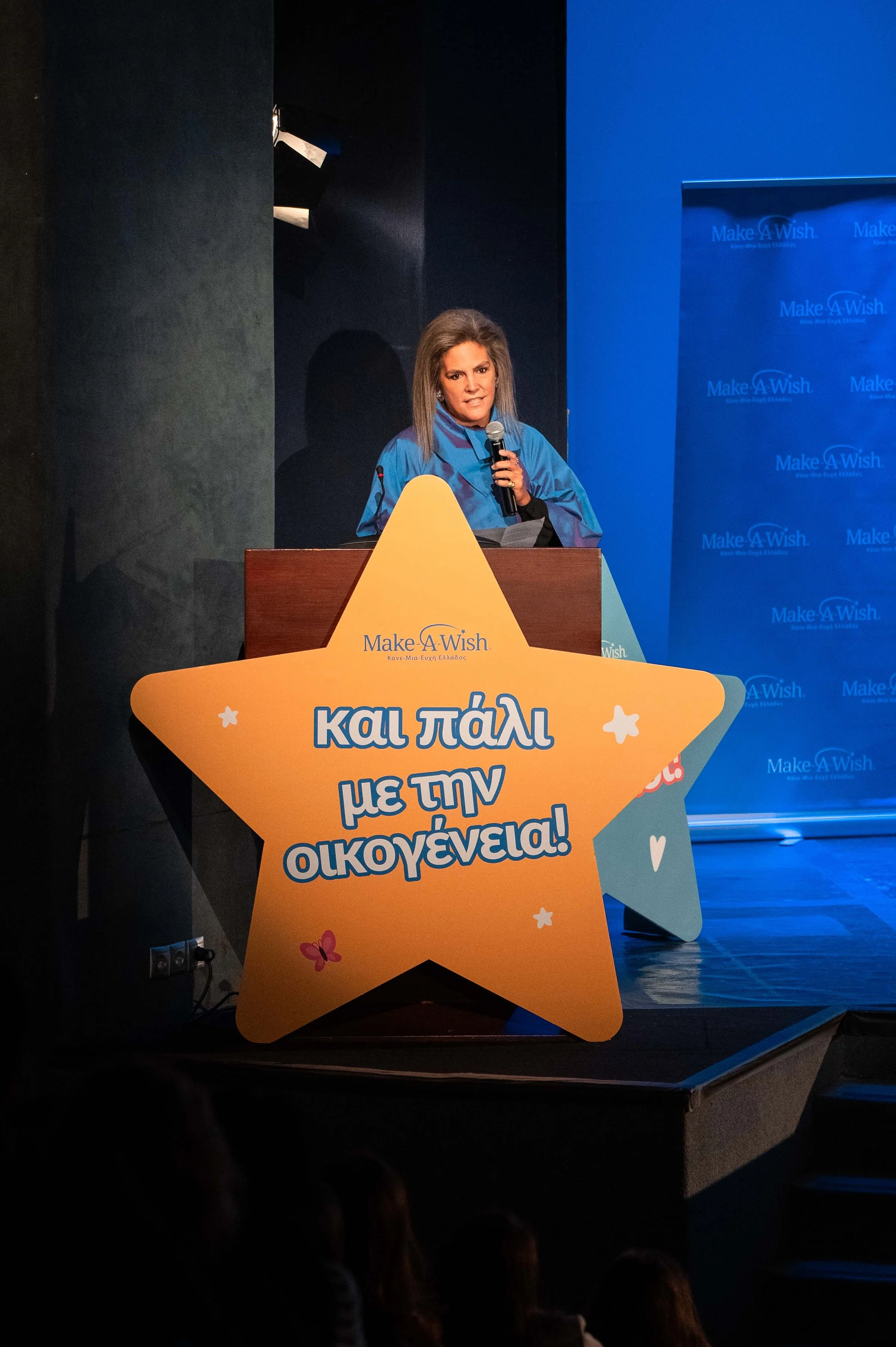 A woman speaking at a podium with a blue Make-A-Wish backdrop behind her. In front of the podium is a large star-shaped sign with Greek text.