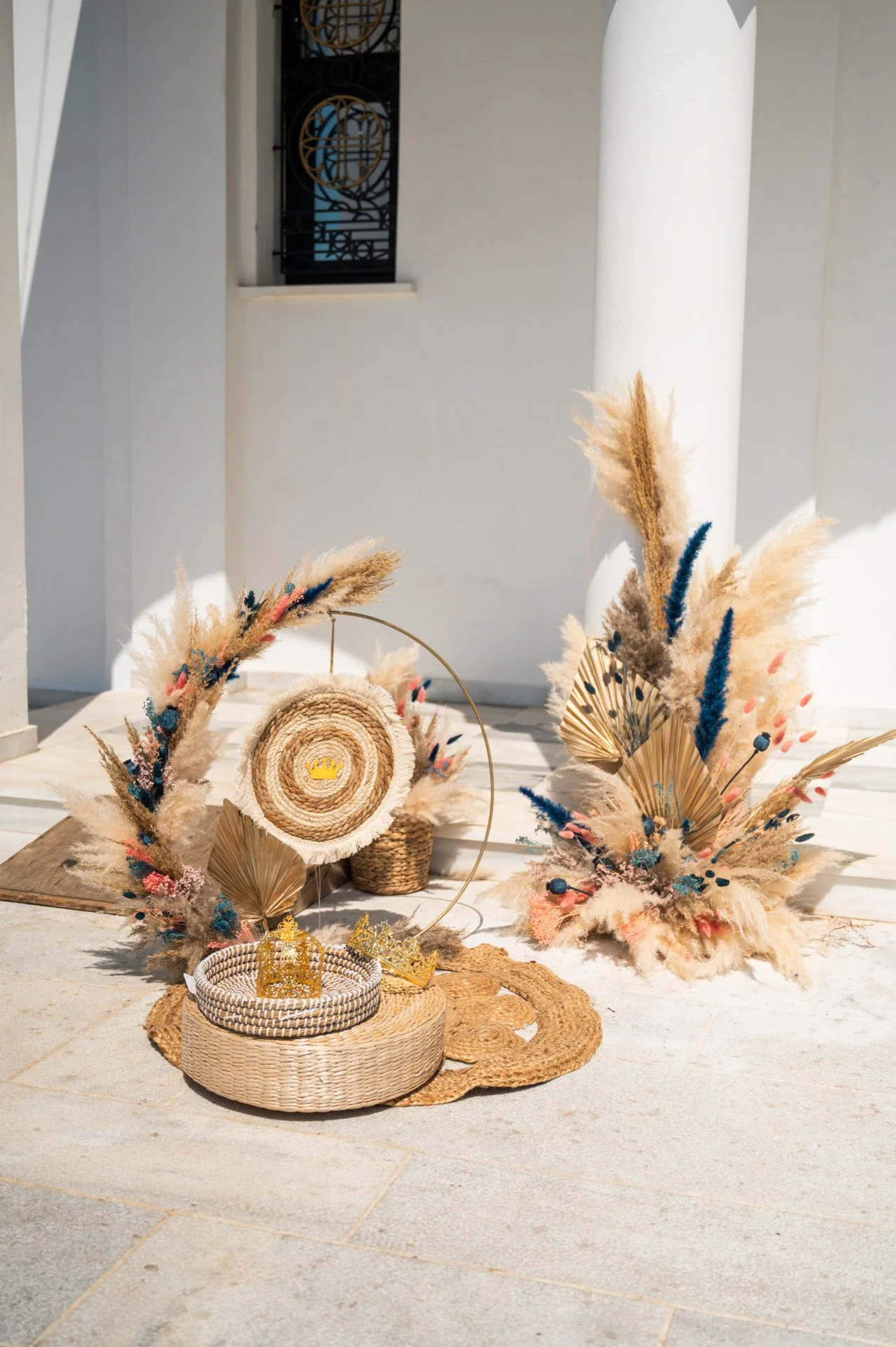 Decorative arrangement with pampas grass, dried leaves, and floral elements in neutral and blue tones, placed on woven baskets and a wooden mat outside a white building.