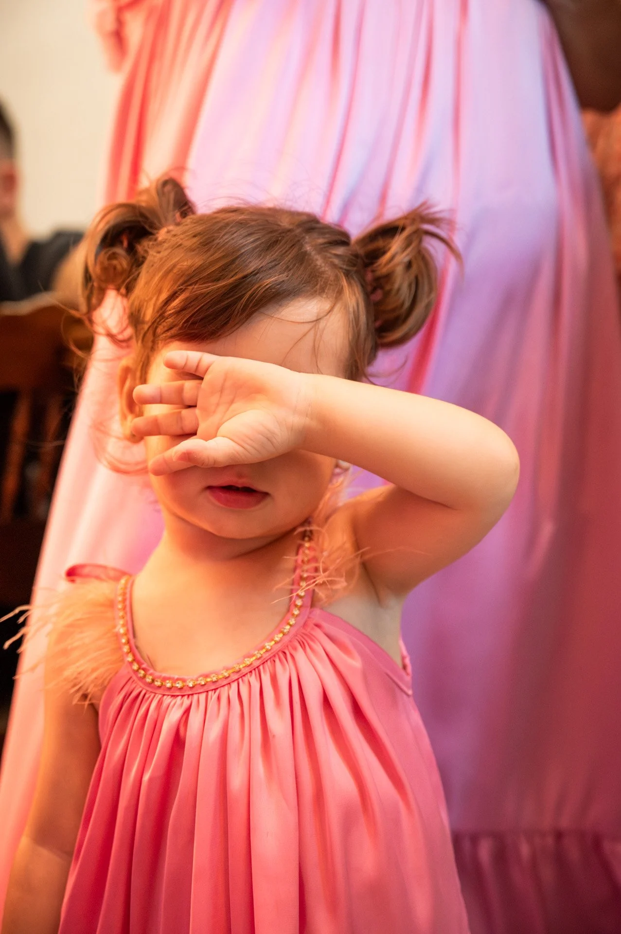 A young girl with brown hair styled in two pigtails, wearing a pink dress with a beaded neckline and feathered trim, covering her eyes with her arm in a crowded room.