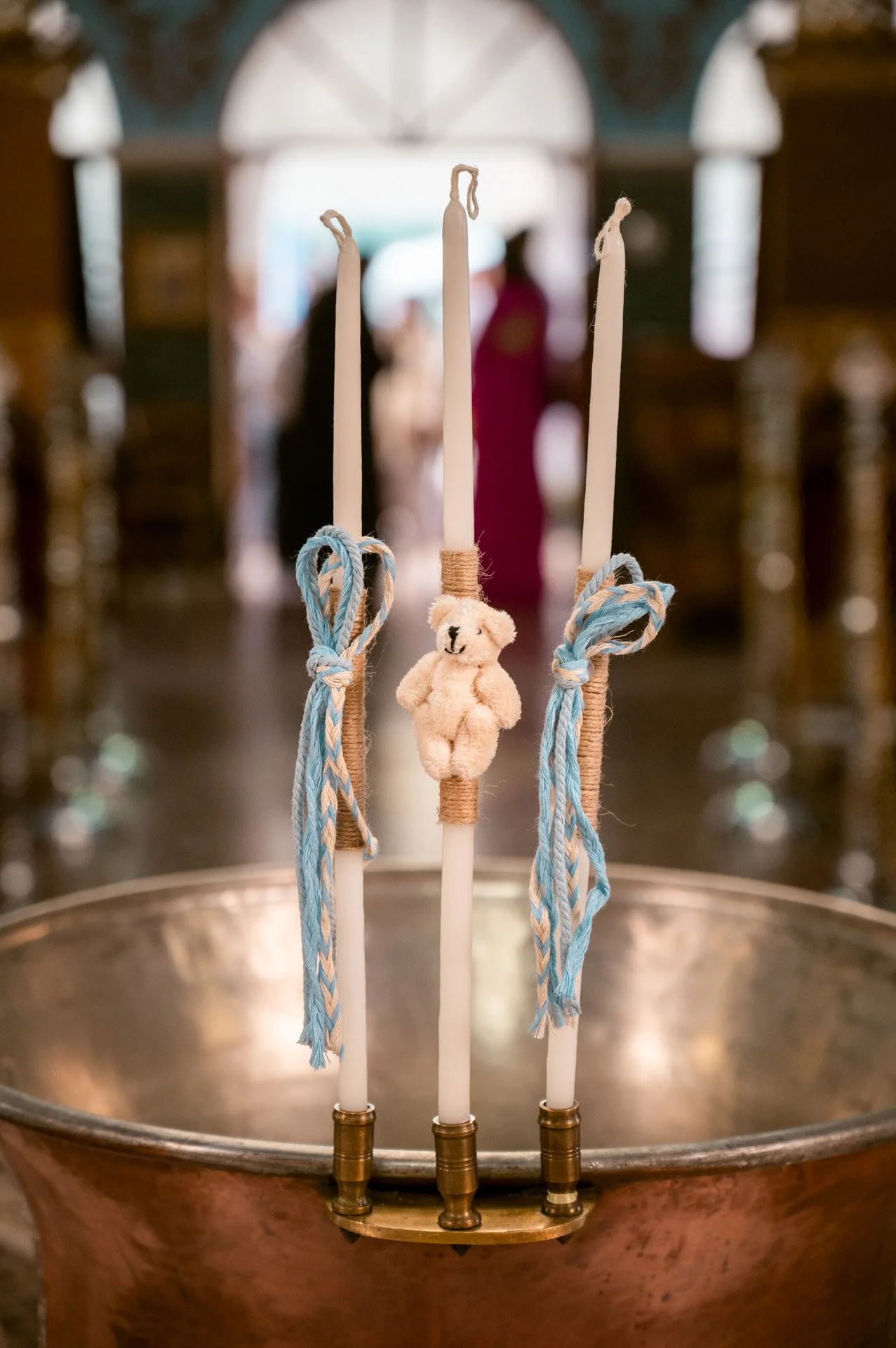 Three white candles, each decorated with a bear clip and tied with blue and white braided ribbons, are arranged in a copper basin for a baptism or church ceremony.