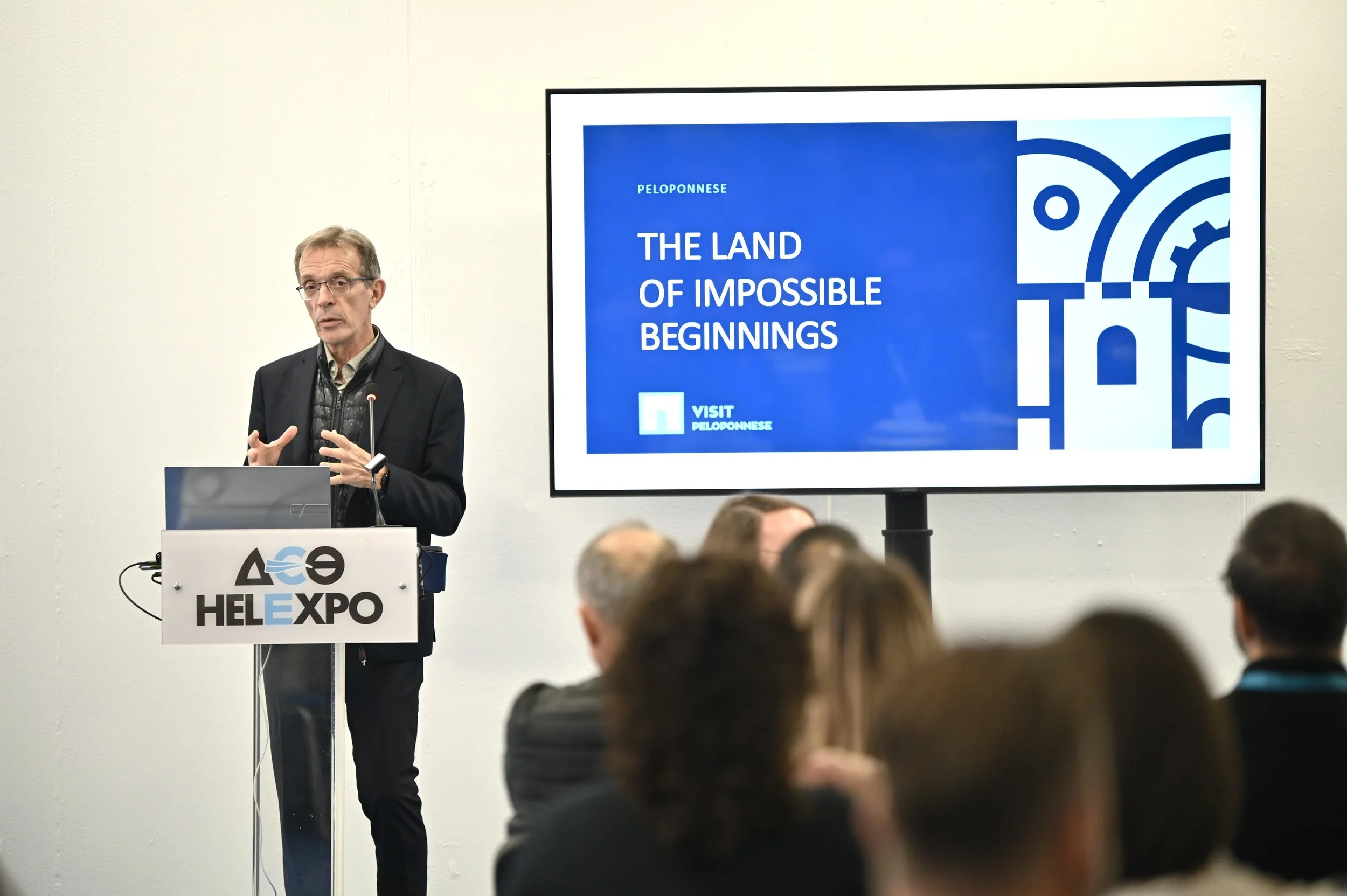 A man in a suit and glasses giving a presentation at a conference with a large screen displaying the text 'Peloponnese, The Land of Impossible Beginnings, Visit Peloponnese.' An audience is seated in front of him, and a podium with the logo 'HEL EXPO