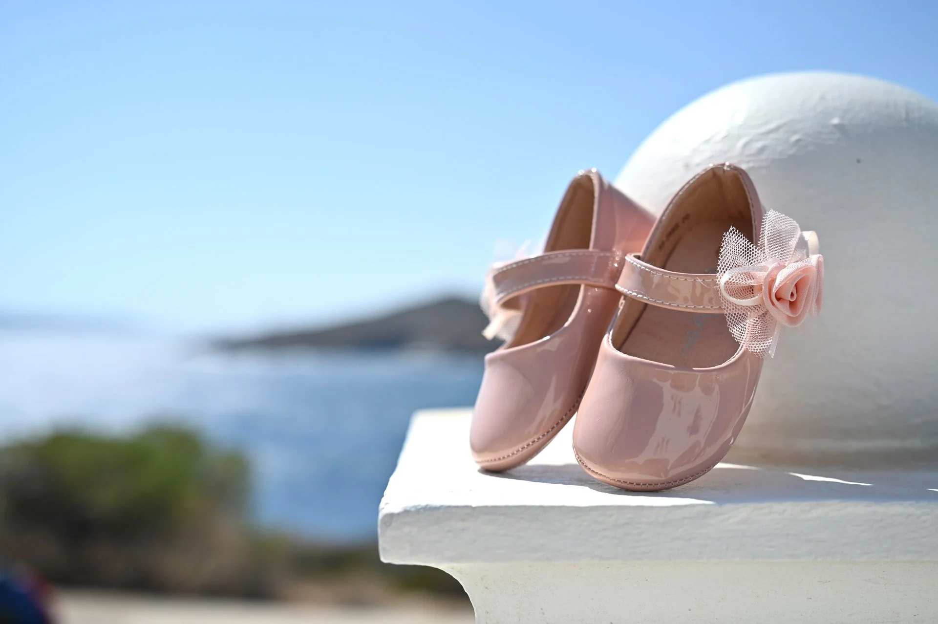 Pink children's shoes with a bow and flower embellishment on the side, placed on a white surface outdoors with a blurred ocean and sky background.