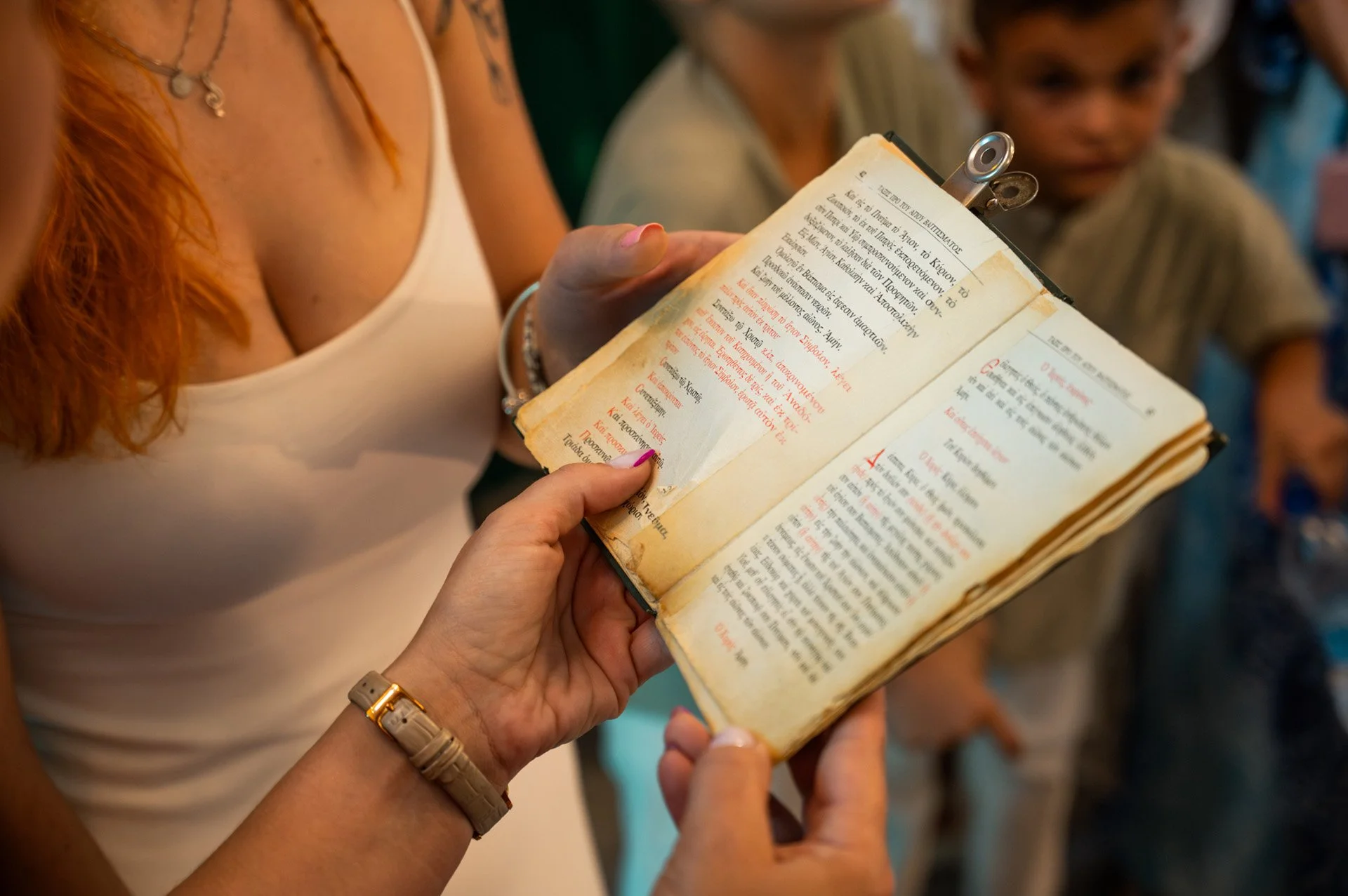 A person with a white tank top and bracelet holding an open ancient book with red and black text, while other people look on.