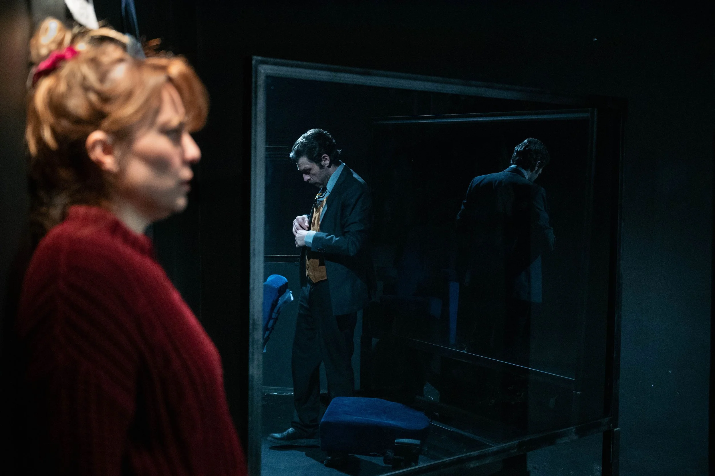 A woman with red hair and a red sweater is in the foreground, out of focus. In the background, a man in a suit and vest is adjusting his tie while looking down, with his reflection visible in a mirror behind him.